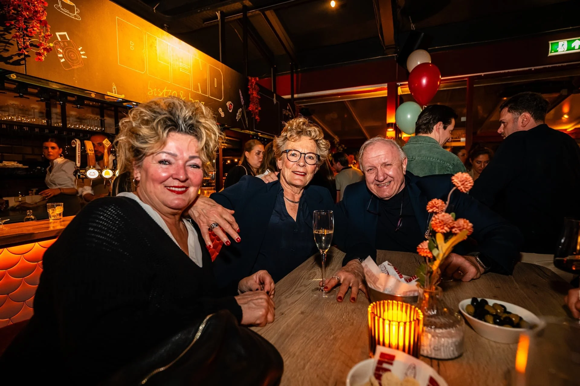 Three older adults, two women and one man, are sitting at a bar or restaurant table, smiling at the camera. The woman on the left has curly blonde hair, and the woman in the center has short, curly blonde hair and glasses. The man on the right has sh