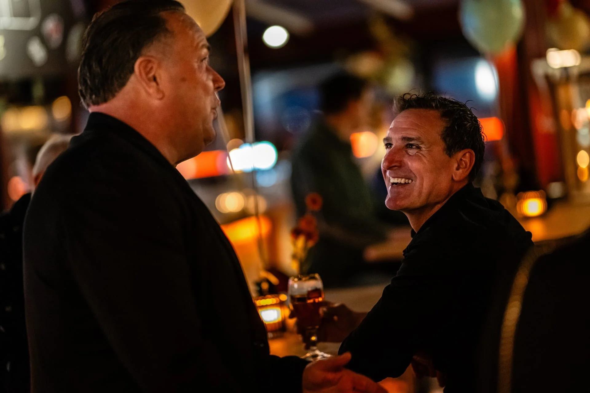 Two men are in a dimly lit bar, smiling and holding hands. The man on the right is seated at the bar, looking up at the man on the left, who is standing. There are blurred lights and bar decorations in the background.