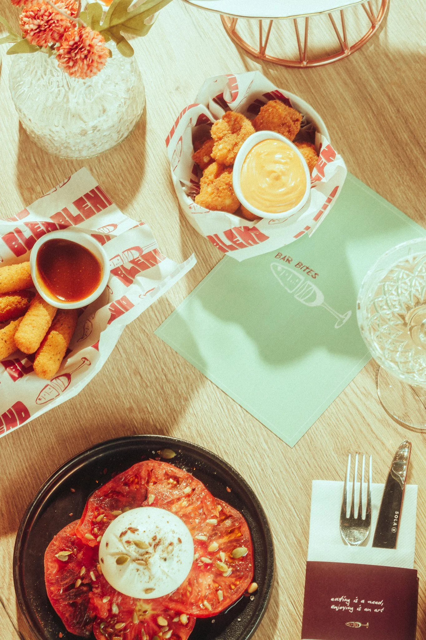 A table with a plate of sliced tomato topped with a soft-boiled egg and seeds, a basket of fried food with dipping sauce, and another basket with fried sticks and sauce, with a glass of water and a flower arrangement in a vase.