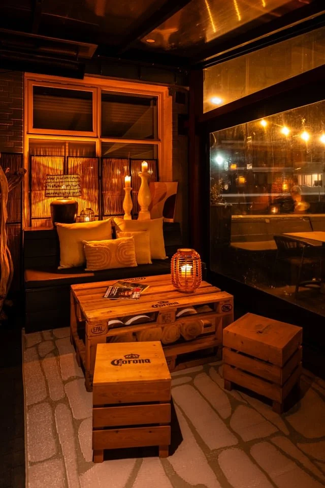 Cozy indoor seating area illuminated with warm lighting, featuring wooden furniture, cushions, and decorative candles, with a large window showcasing a night street view.