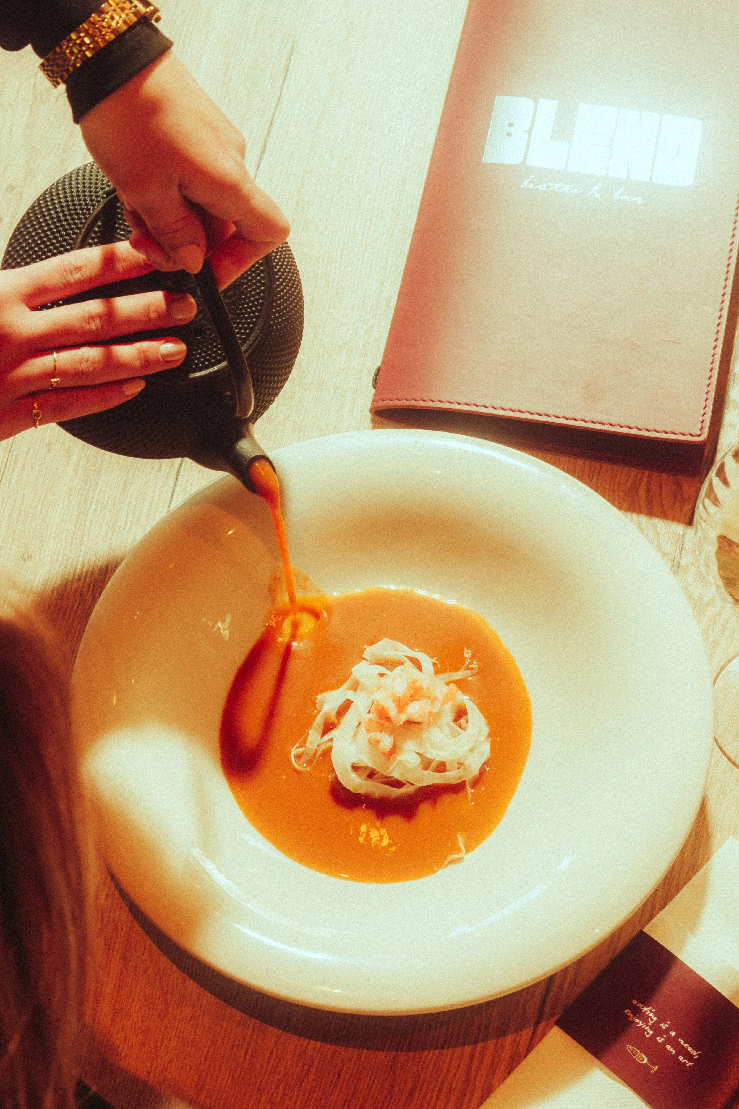 Pouring orange soup or sauce onto a plate with shredded cheese or toppings, on a wooden table.