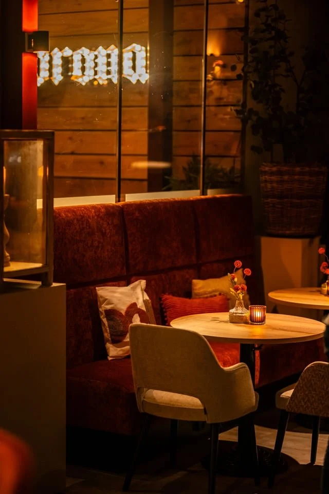 A cozy restaurant interior with dim lighting, a plush red velvet banquette, a small round wooden table with a candle and small flower vase, and upholstered chairs, with wooden wall panels and a large potted plant in the background.