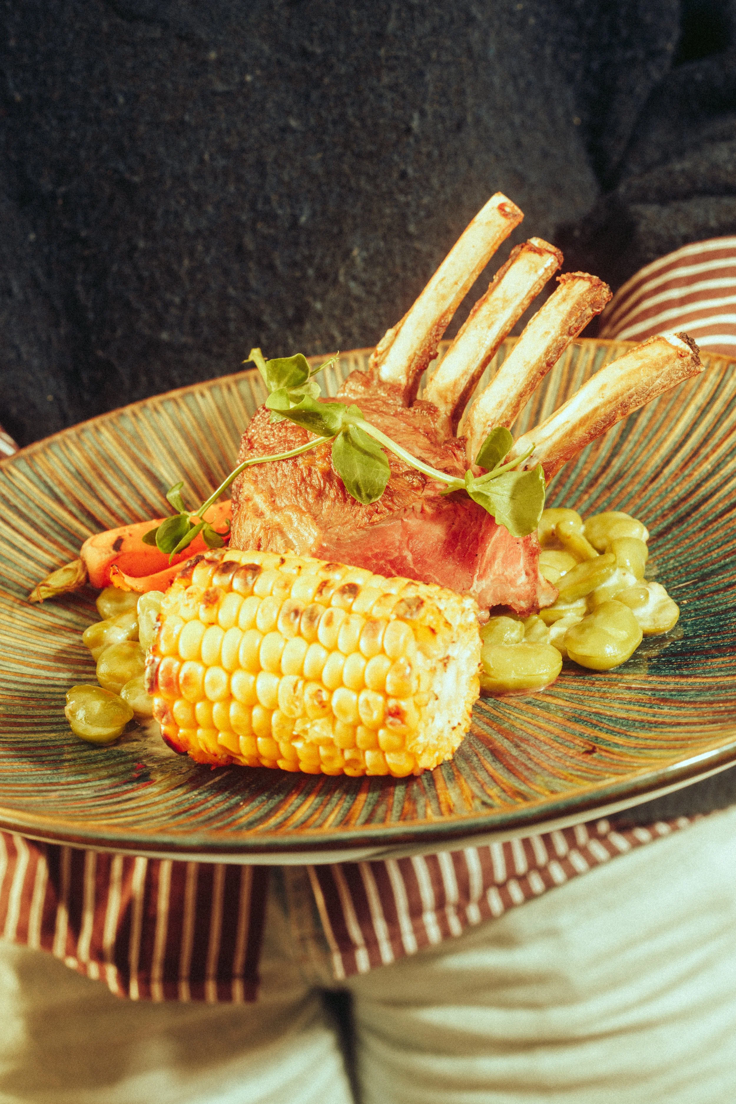 A plate of food featuring a rack of lamb chops, corn on the cob, and some green beans, garnished with a sprig of herbs.