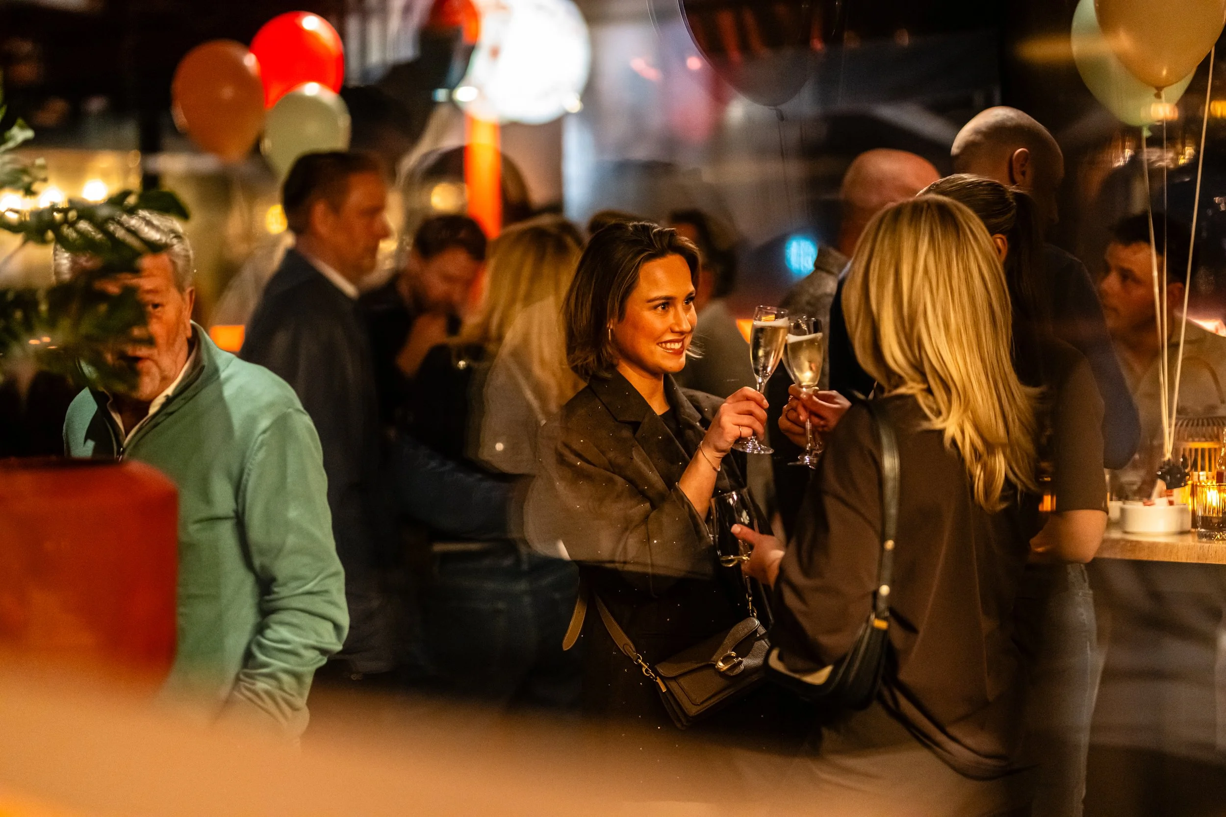 People celebrating with champagne glasses at a bar or party, with balloons and warm lighting in the background.