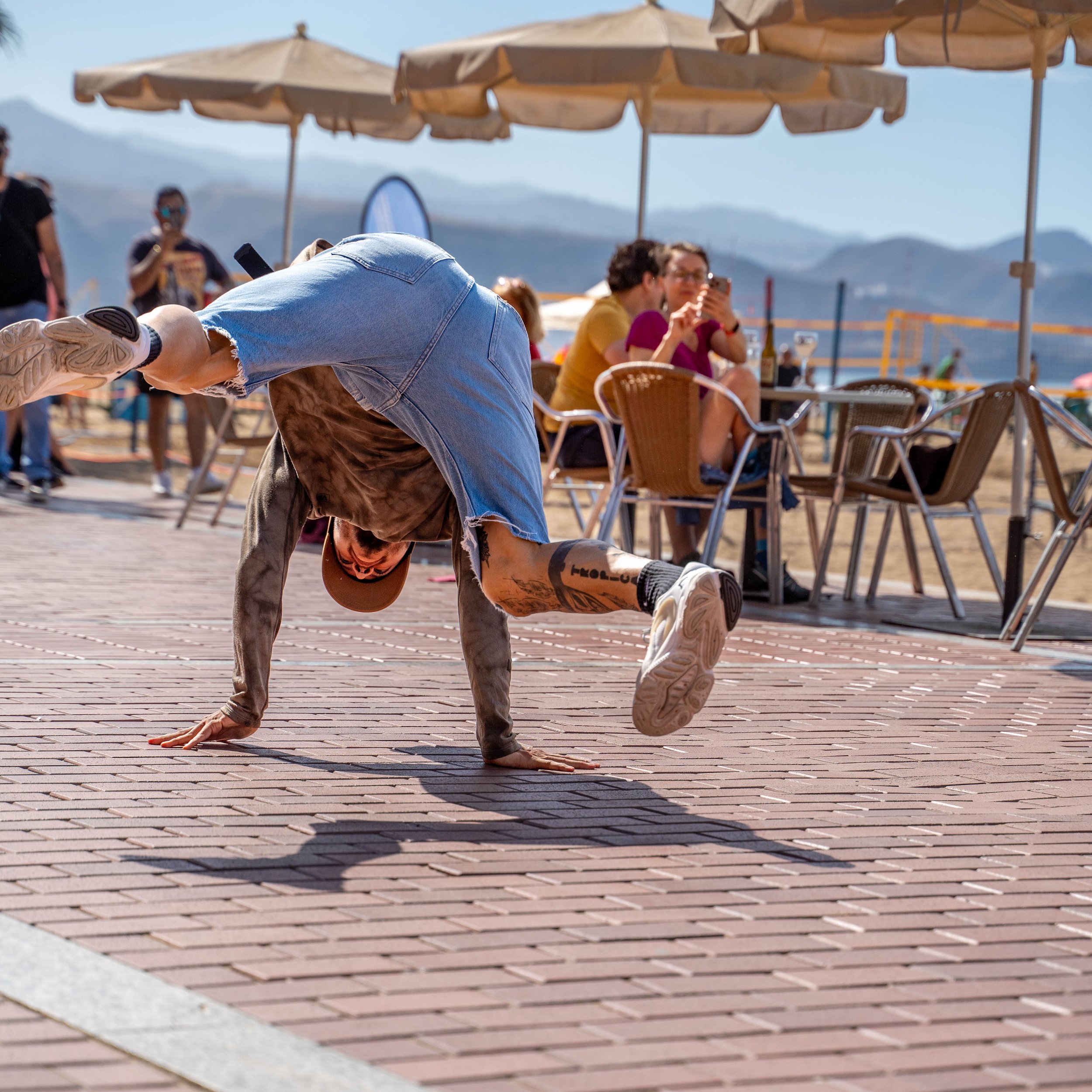A person performing a handstand on a brick pavement at an outdoor beachside cafe with umbrellas, while people at the cafe sit at tables, some taking photos.