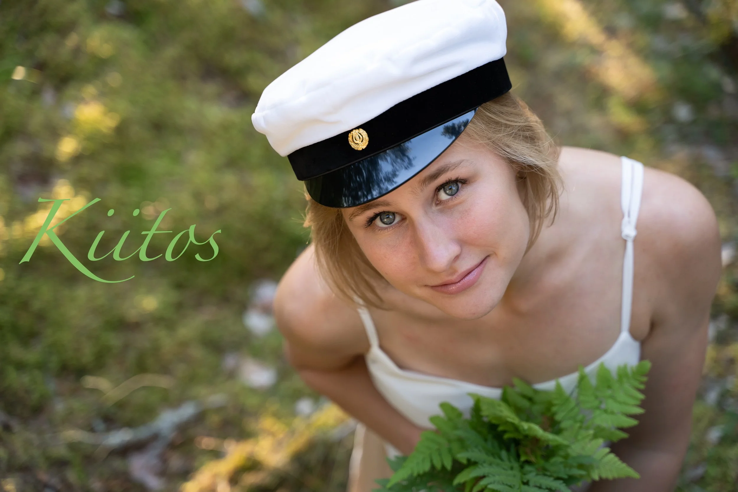A young woman with light brown hair and blue eyes, wearing a white dress and a white and black captain's hat, holding a green fern, outdoors with blurred greenery in the background.