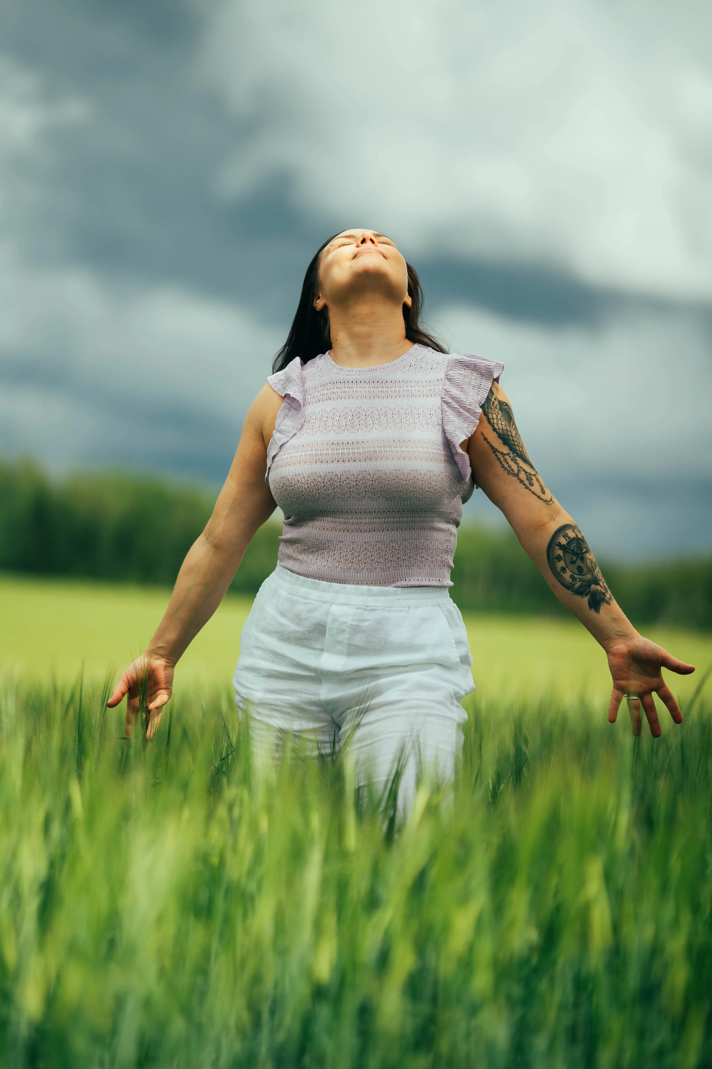 A woman standing in a lush green field with her arms outstretched and head tilted back, enjoying the outdoors under dark, cloudy sky.