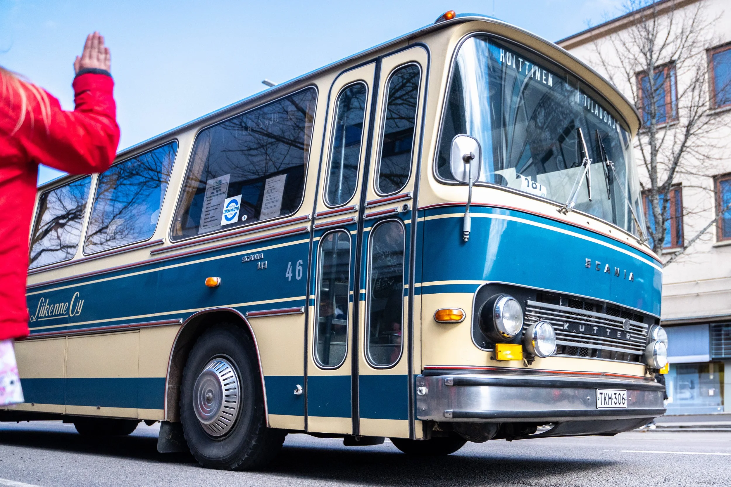 Vintage blue and cream bus parked on a street, with a person in red jacket raising their hand nearby.
