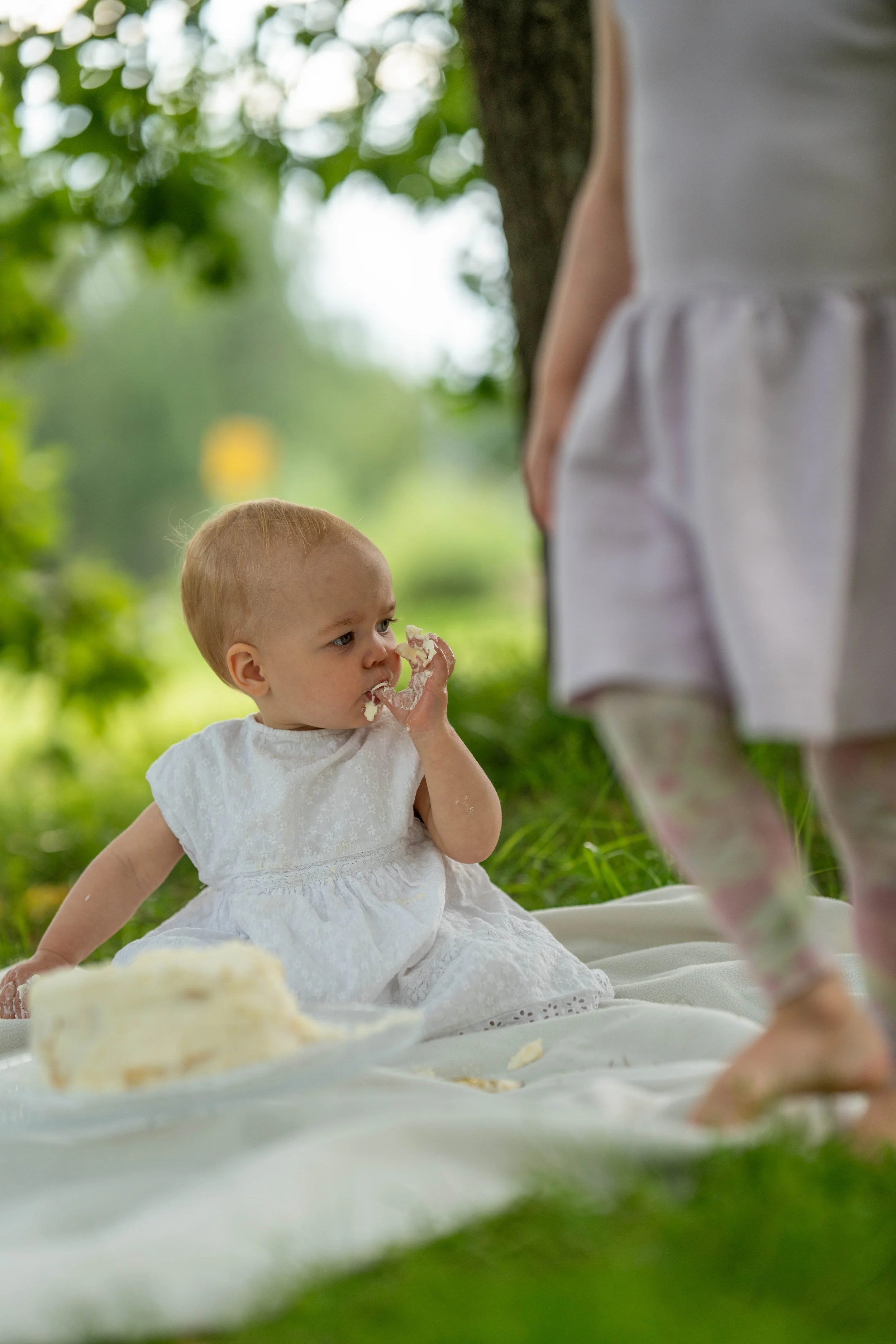 A young child in a white dress sitting on a white blanket outdoors, eating cake, with another person standing nearby.