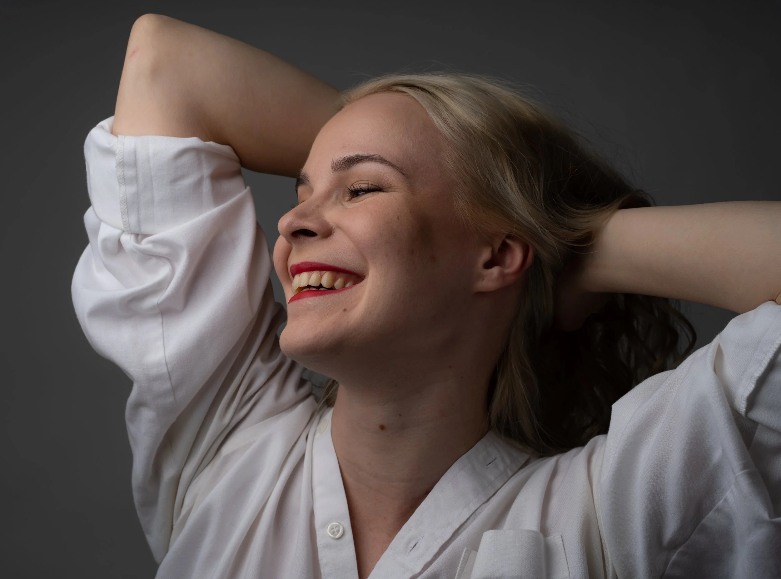 A smiling woman with blonde hair in a white shirt, touching her head with her hands, against a dark background.