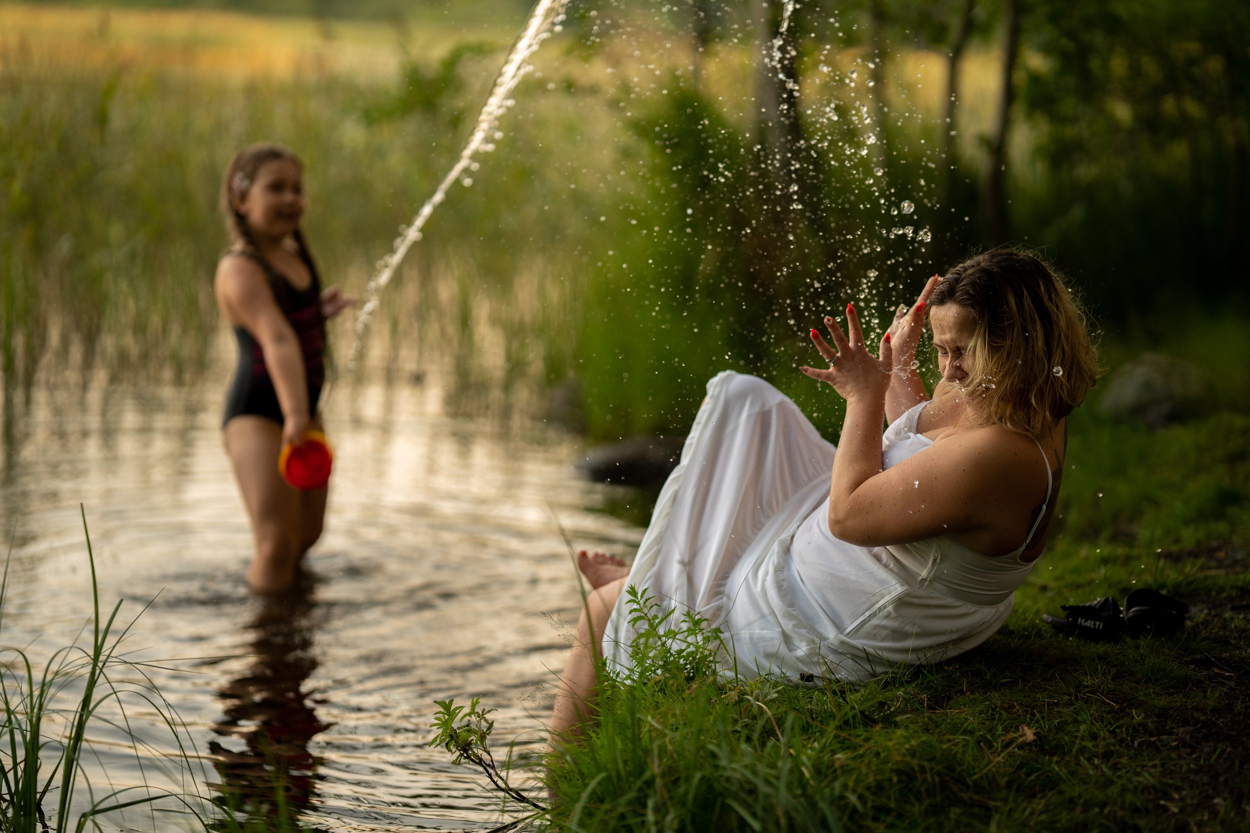 A woman in a white dress sitting by a lake, splashing water on her face as a young girl in a swimsuit stands in the water holding a toy watering can and spraying water towards her. The background features green trees and blurred scenery.