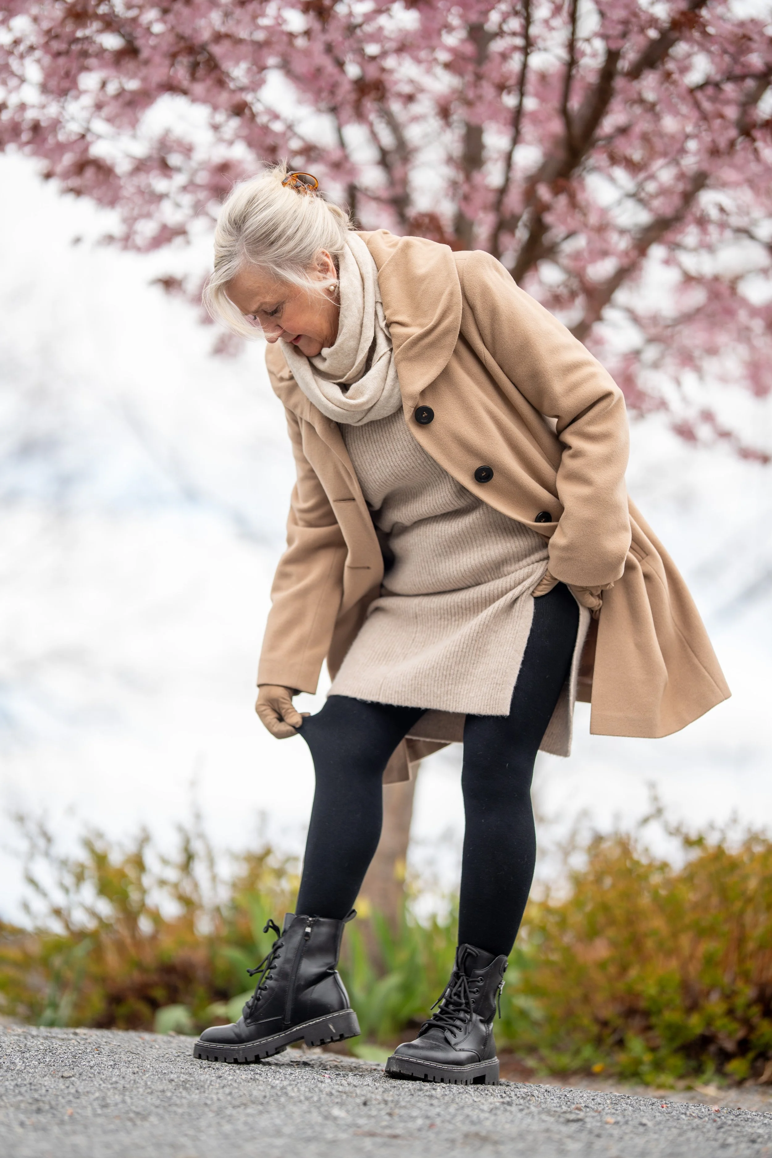 An elderly woman dressed in a beige coat, sweater, and scarf looking down at her black boots, with pink blossoms on a tree in the background.