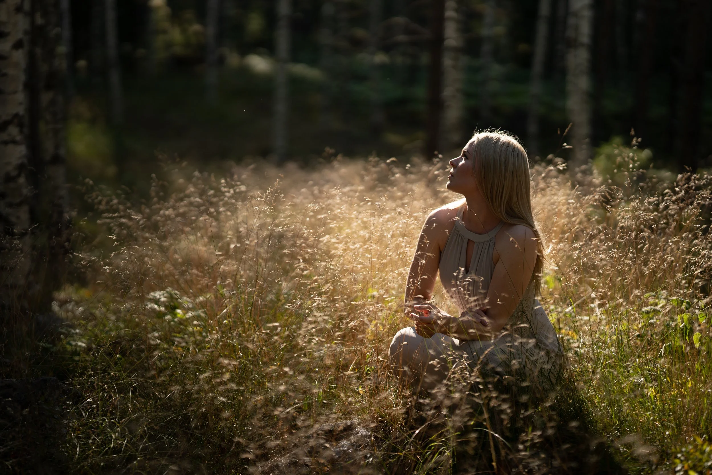A woman with long blonde hair in a beige dress sitting in a sunlit grassy area in a forest, looking upwards.