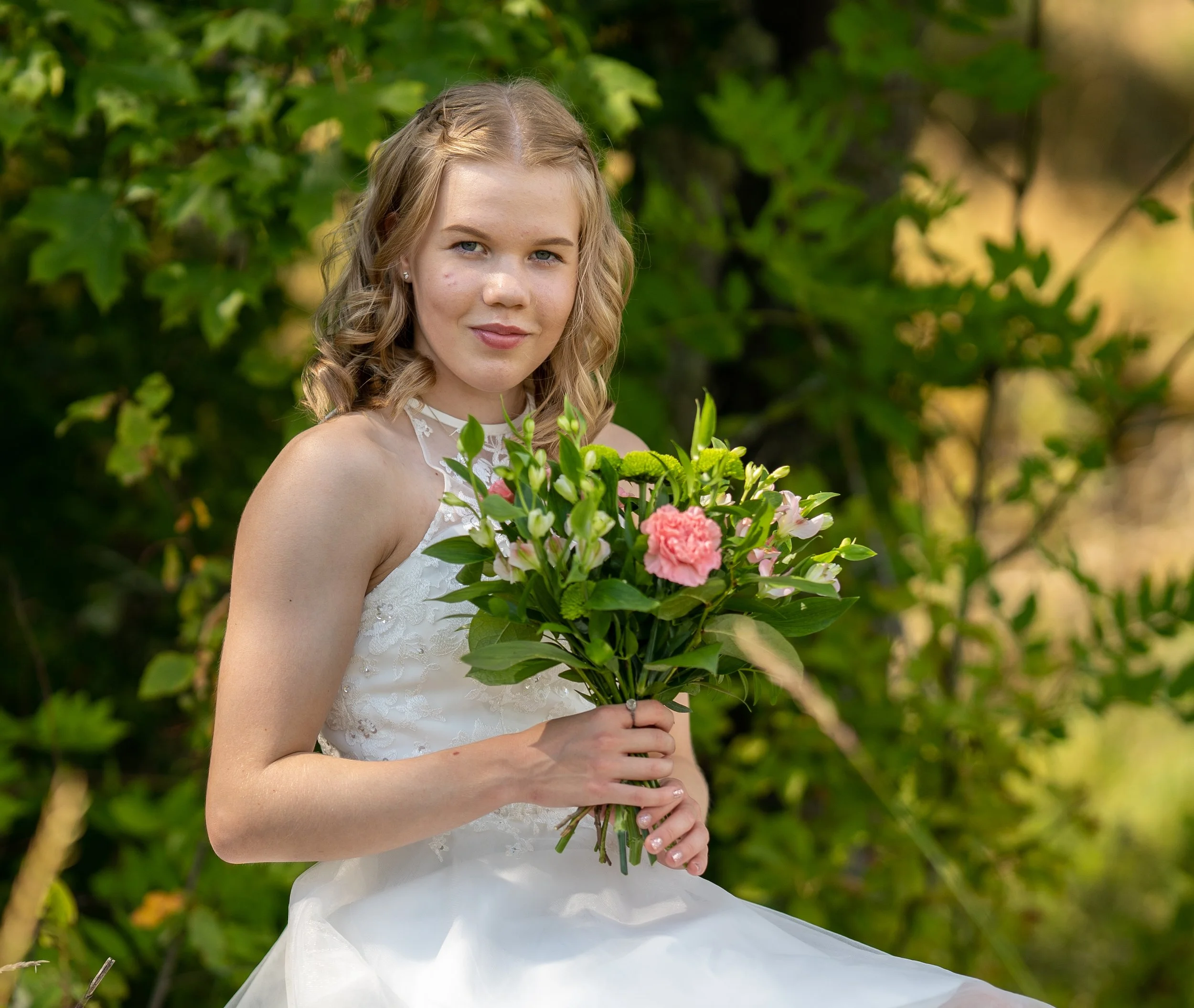 Young woman in a white dress holding a bouquet of pink and white flowers outdoors with green foliage in the background.