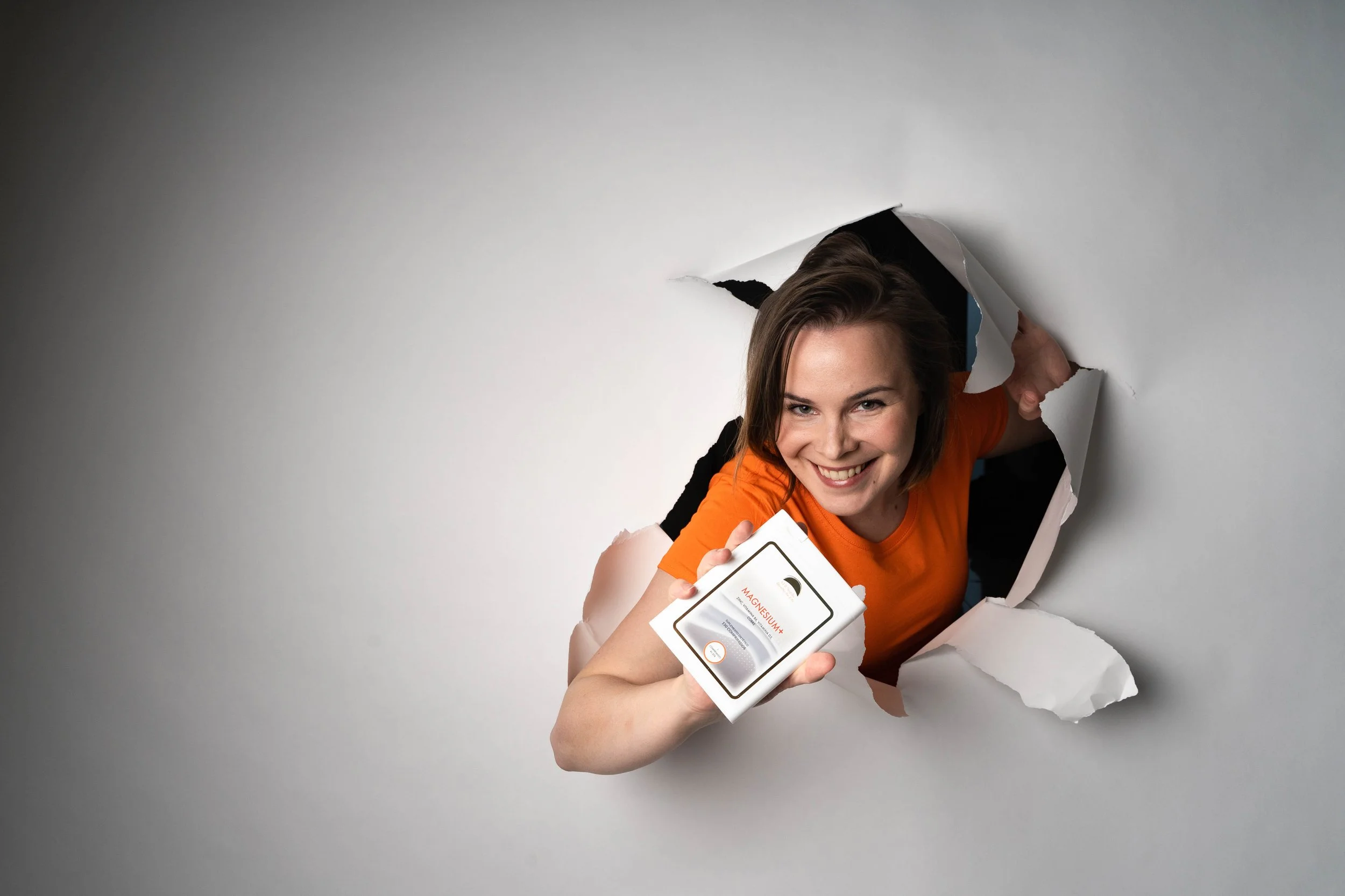 Smiling woman in an orange shirt breaking through a white wall, holding a box of magnesium supplements.