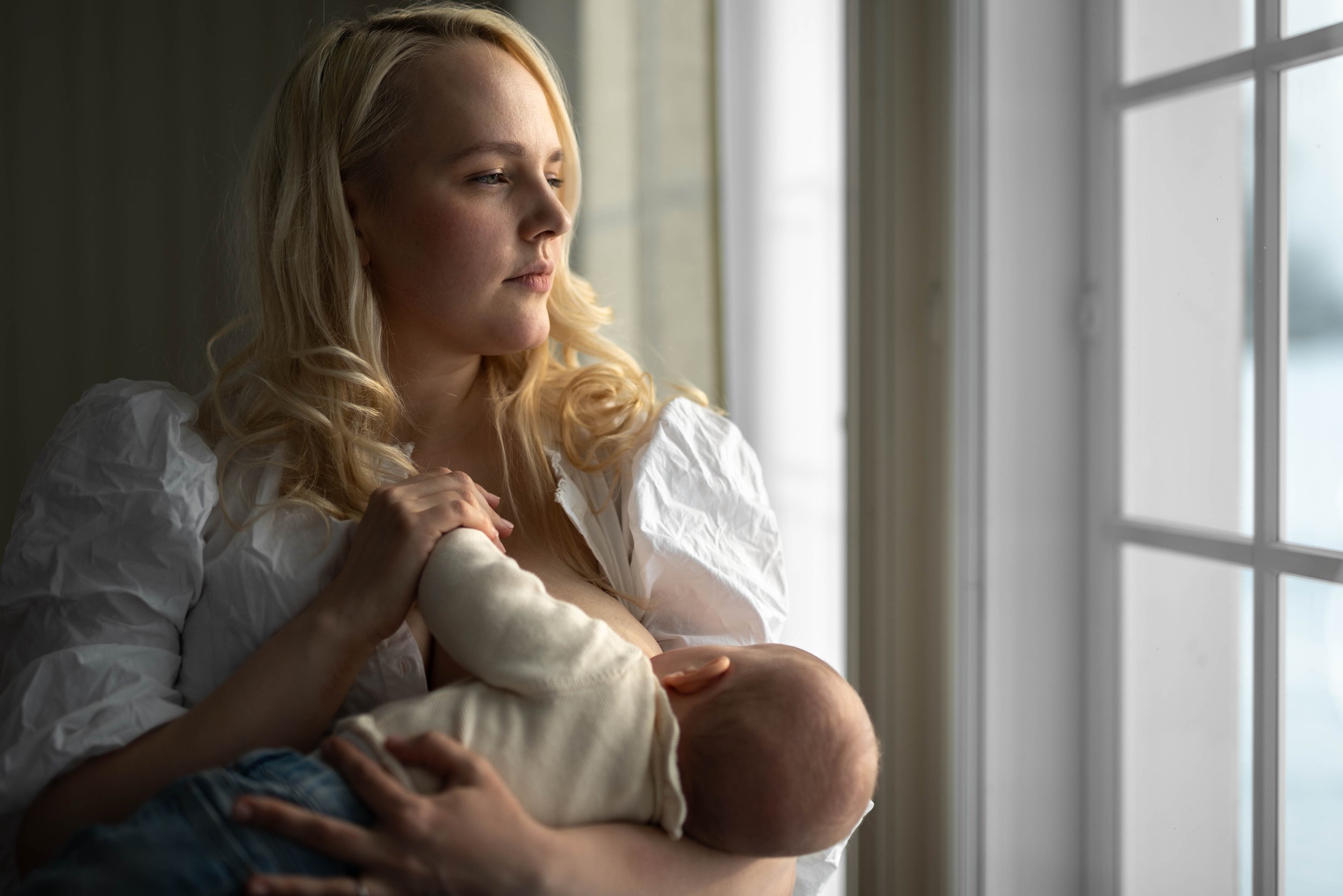 A woman with blonde hair breastfeeding a baby while looking out a window.