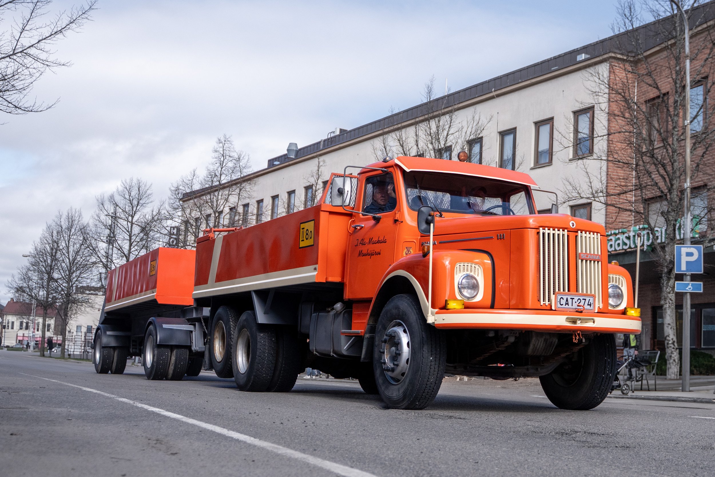 An orange vintage Scania tanker truck driving down a city street with buildings and leafless trees in the background.