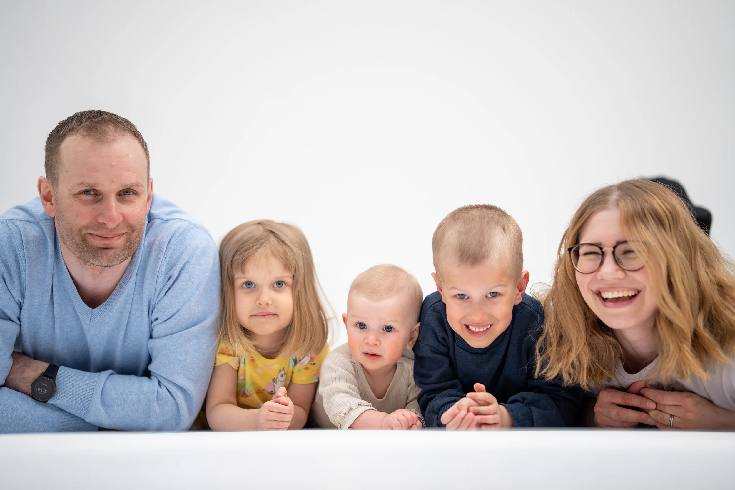 Family laying on the floor in a row, smiling and looking at the camera, against a plain white background.