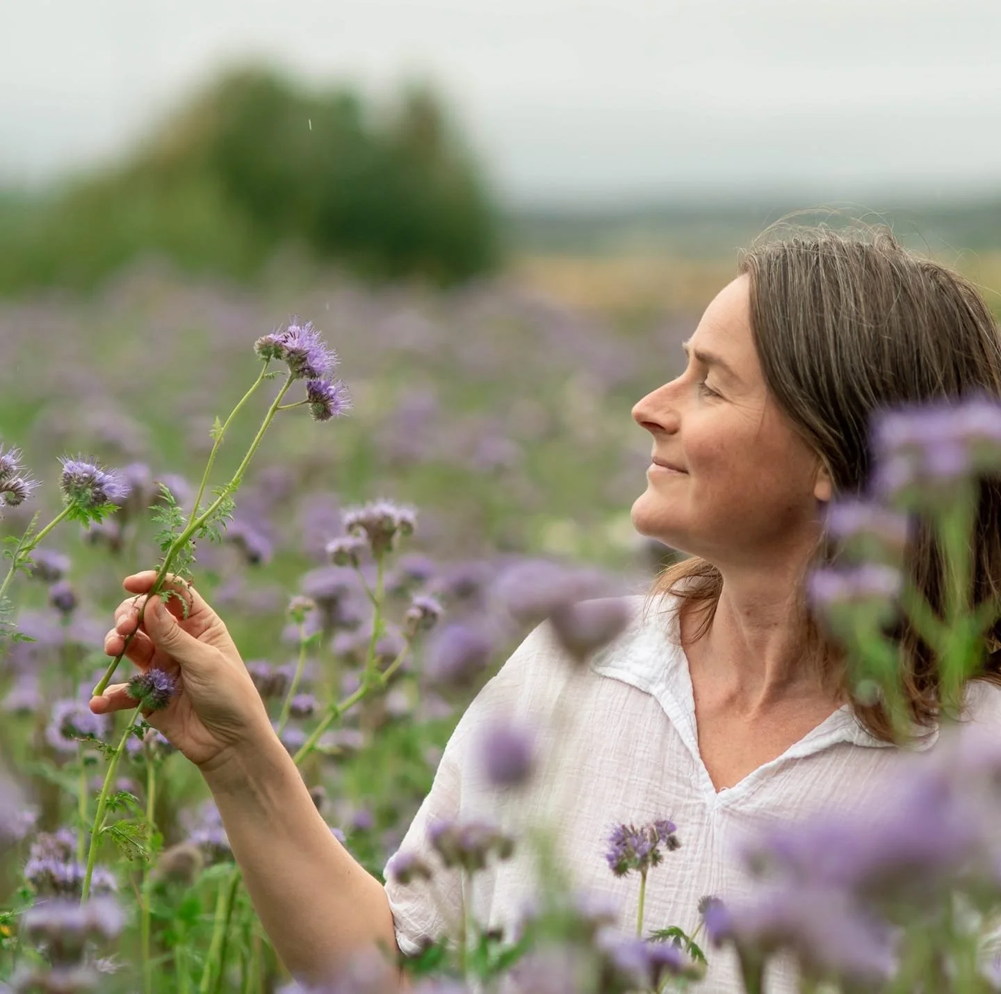 A woman smiling in a field of purple flowers, holding a flower in her hand and enjoying the outdoors.