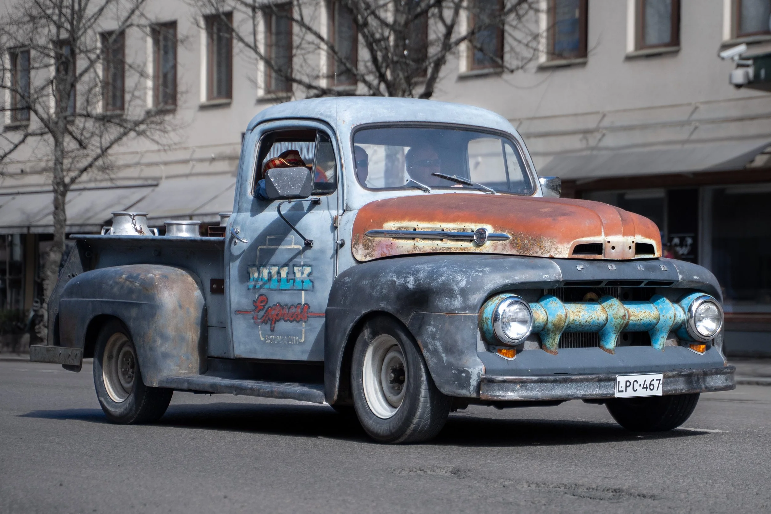 A vintage Ford pickup truck with a rusty, weathered exterior and patched-up paint, parked on a city street.