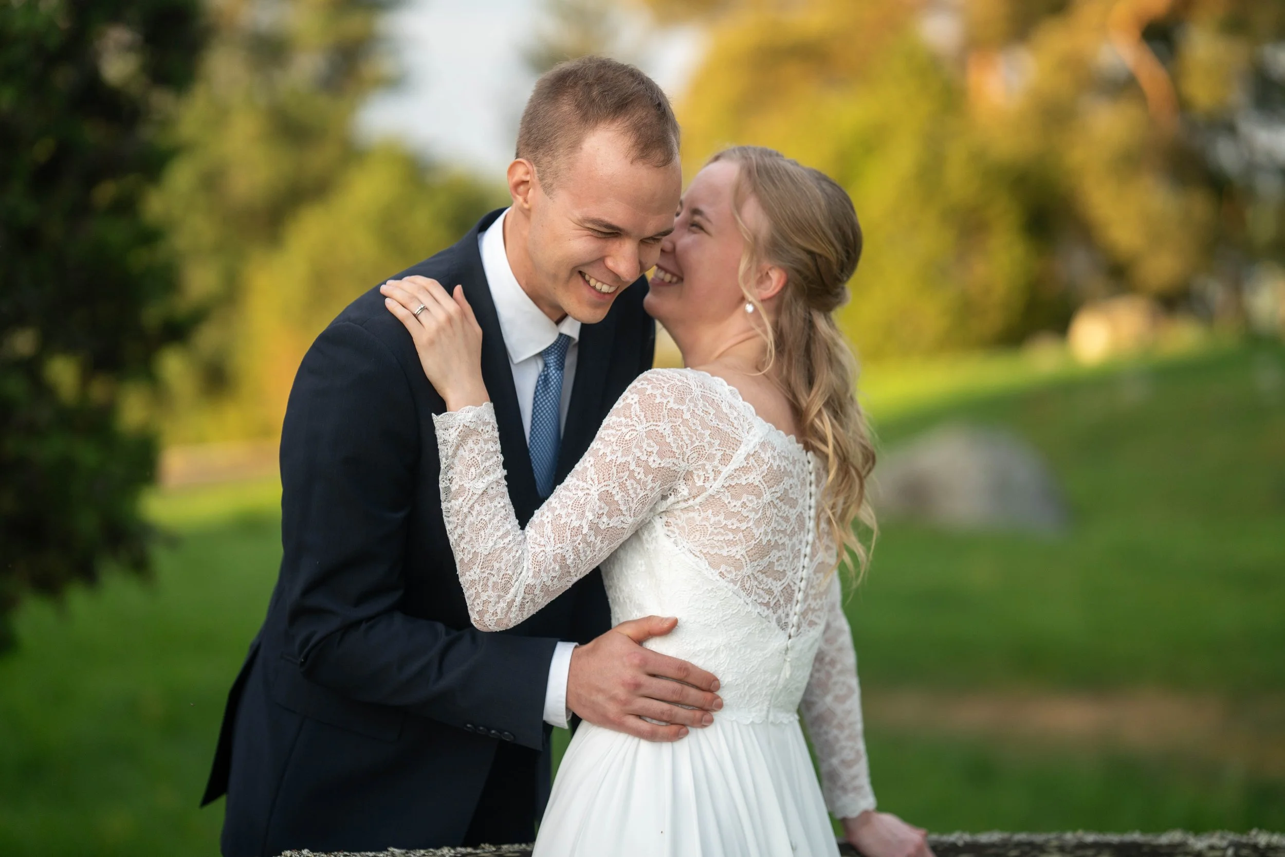 A smiling bride and groom share a joyful moment outdoors, with the bride in a lace wedding dress and the groom in a dark suit, surrounded by greenery and autumn foliage.