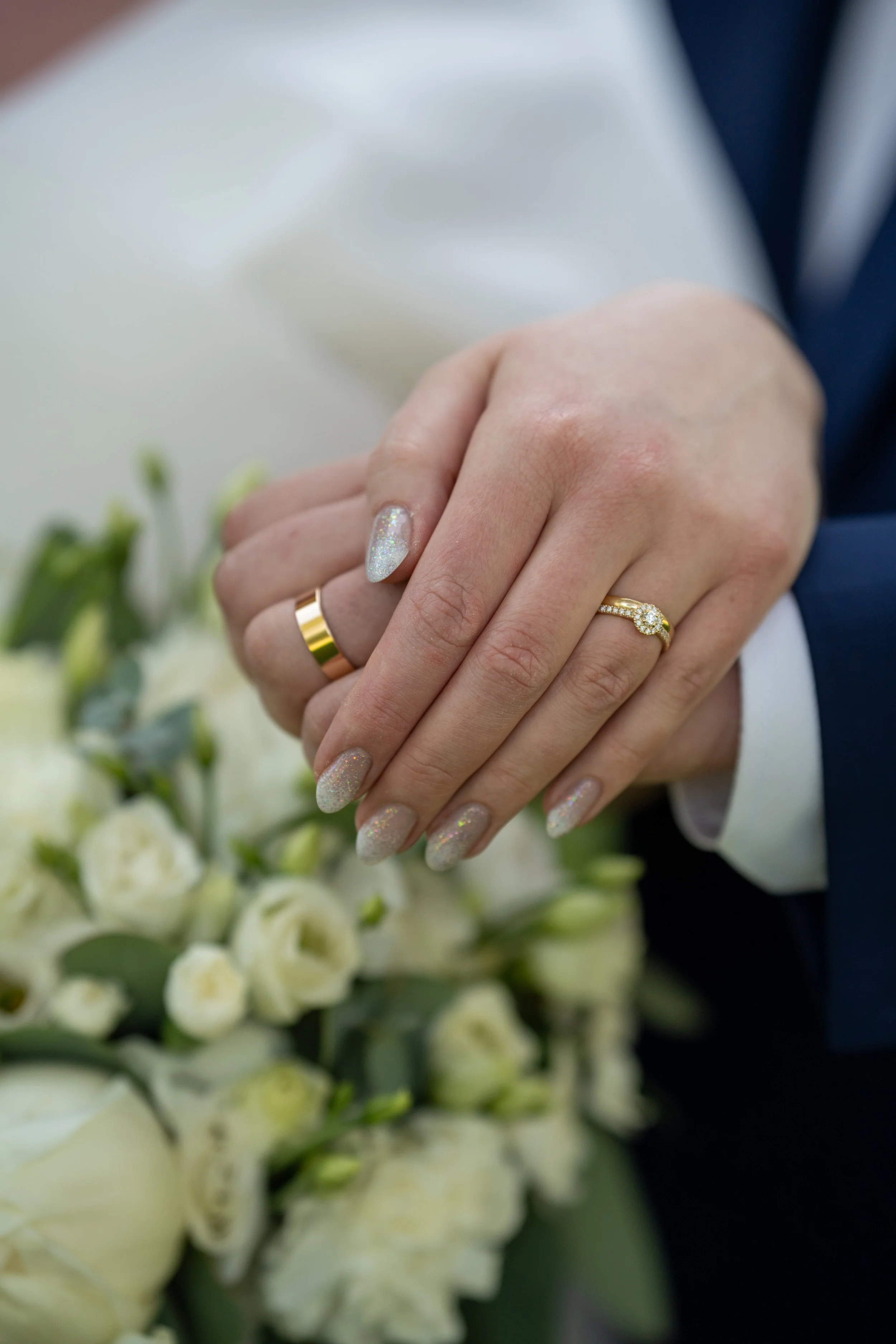 Close-up of a couple's hands with wedding rings, holding a bouquet of white flowers, wearing formal attire.