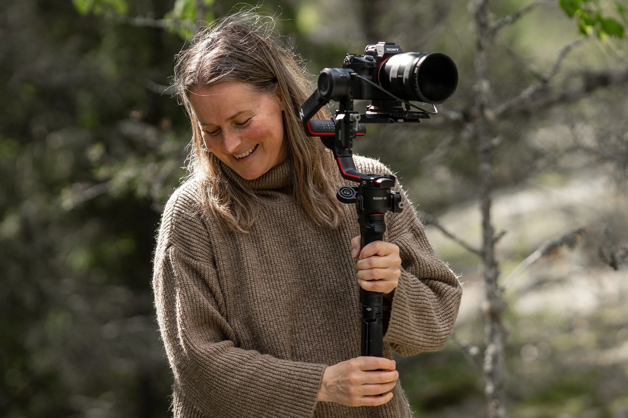 A woman outdoors holding a camera stabilizer with a large lens attached, smiling and looking down, surrounded by trees and natural scenery.