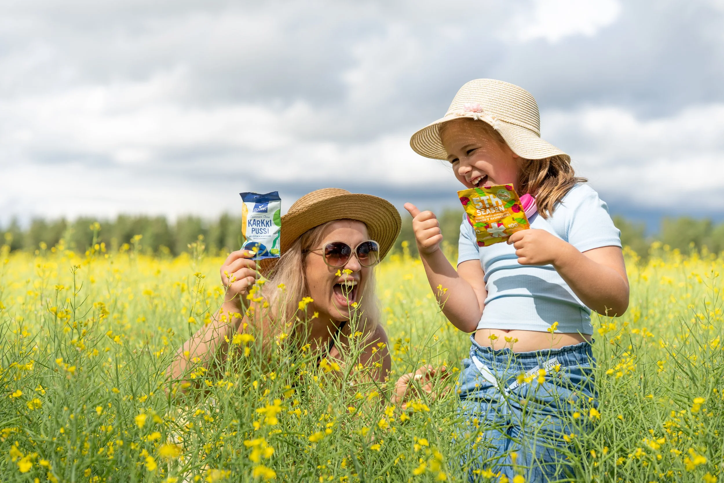 A woman and a young girl in a yellow flower field, both wearing summer hats and sunglasses, laughing and playing with snacks on a cloudy day.