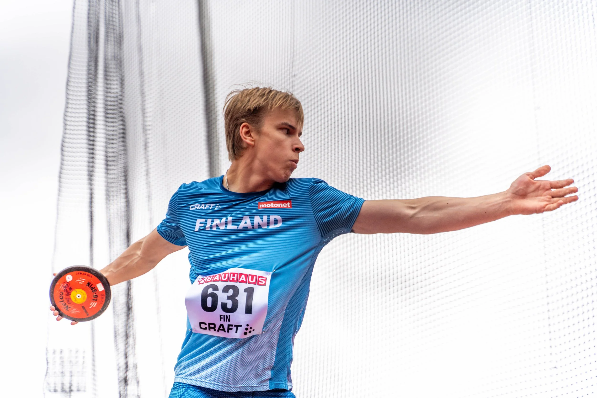 A male athlete wearing a blue Finland jersey and running bib number 631 throws a discus outdoors against a cloudy sky.