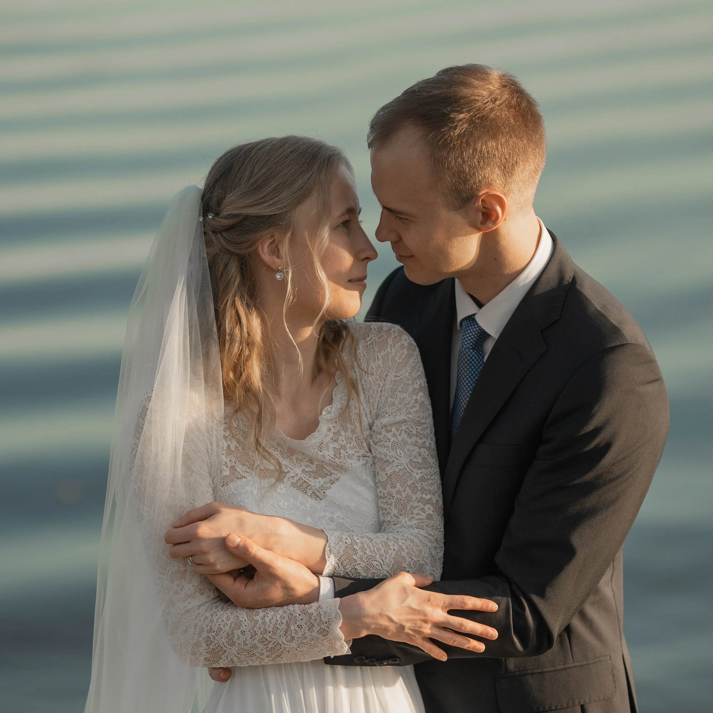 A bride and groom are standing close together near water, gazing into each other's eyes, with the bride wearing a lace wedding dress and veil, and the groom in a black suit and white shirt.