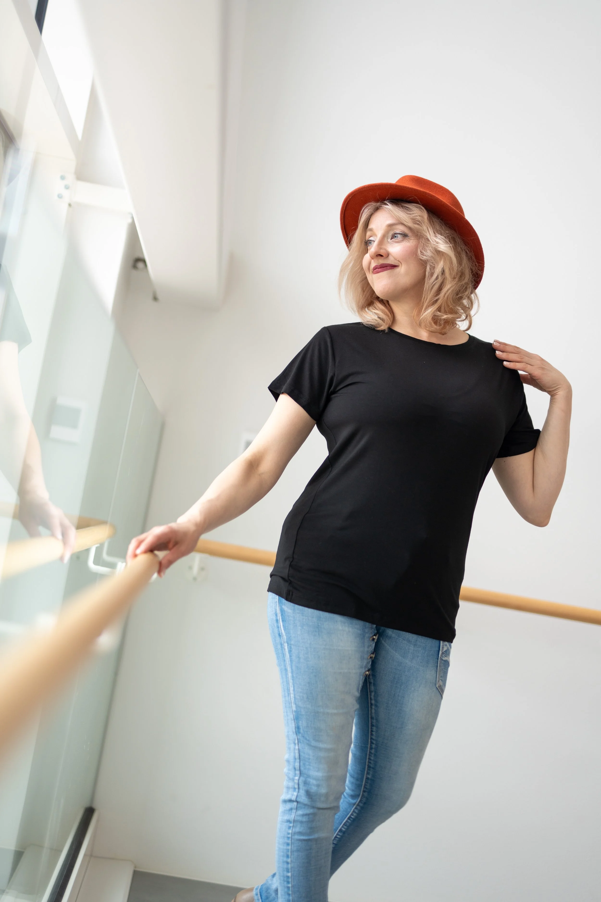 A woman wearing a red hat, black t-shirt, and blue jeans standing at a ballet barre, touching her shoulder with one hand, and looking confidently to the side.