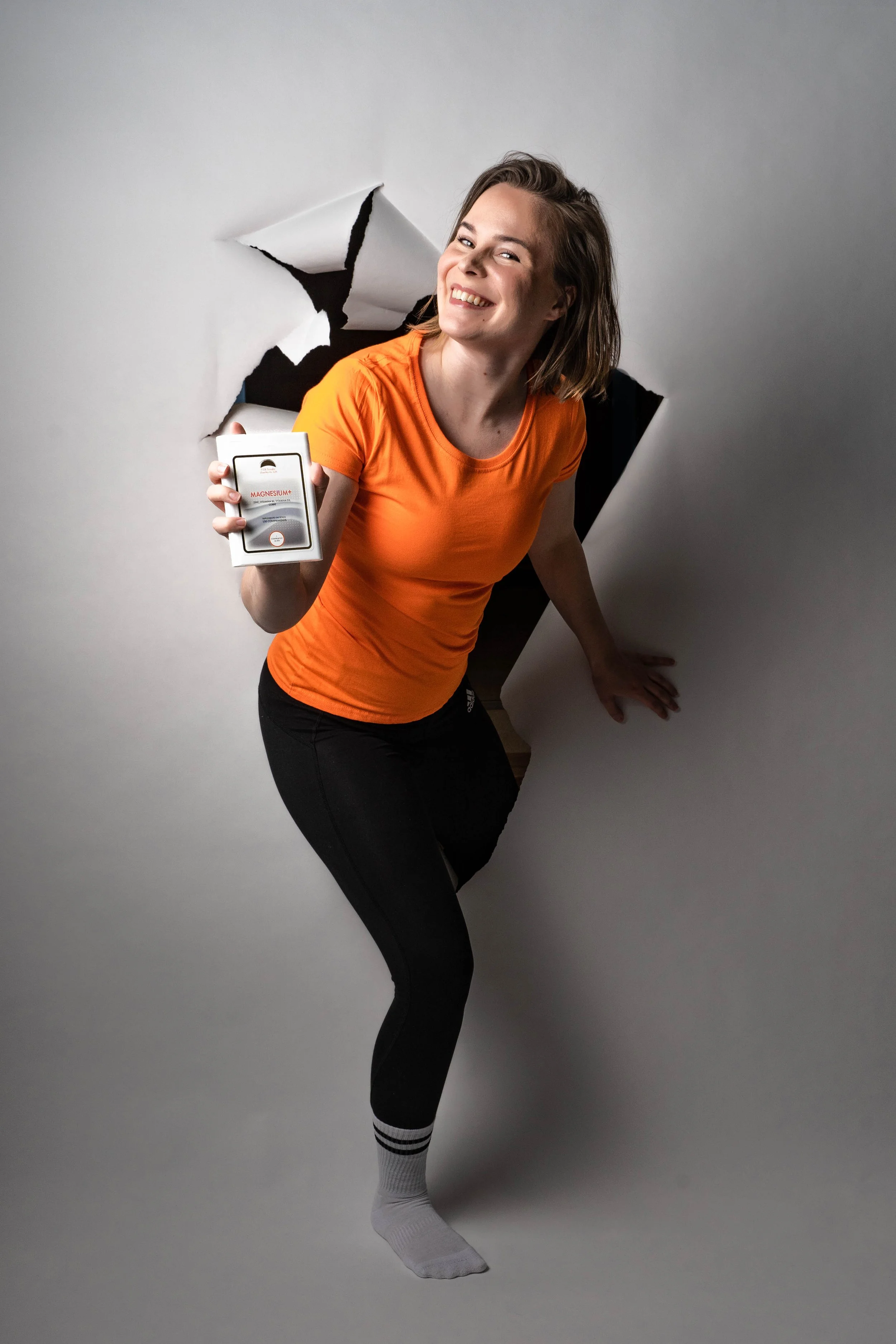 Woman with short brown hair smiling, emerging through a large hole in a white wall, holding a box labeled "Magnesium".