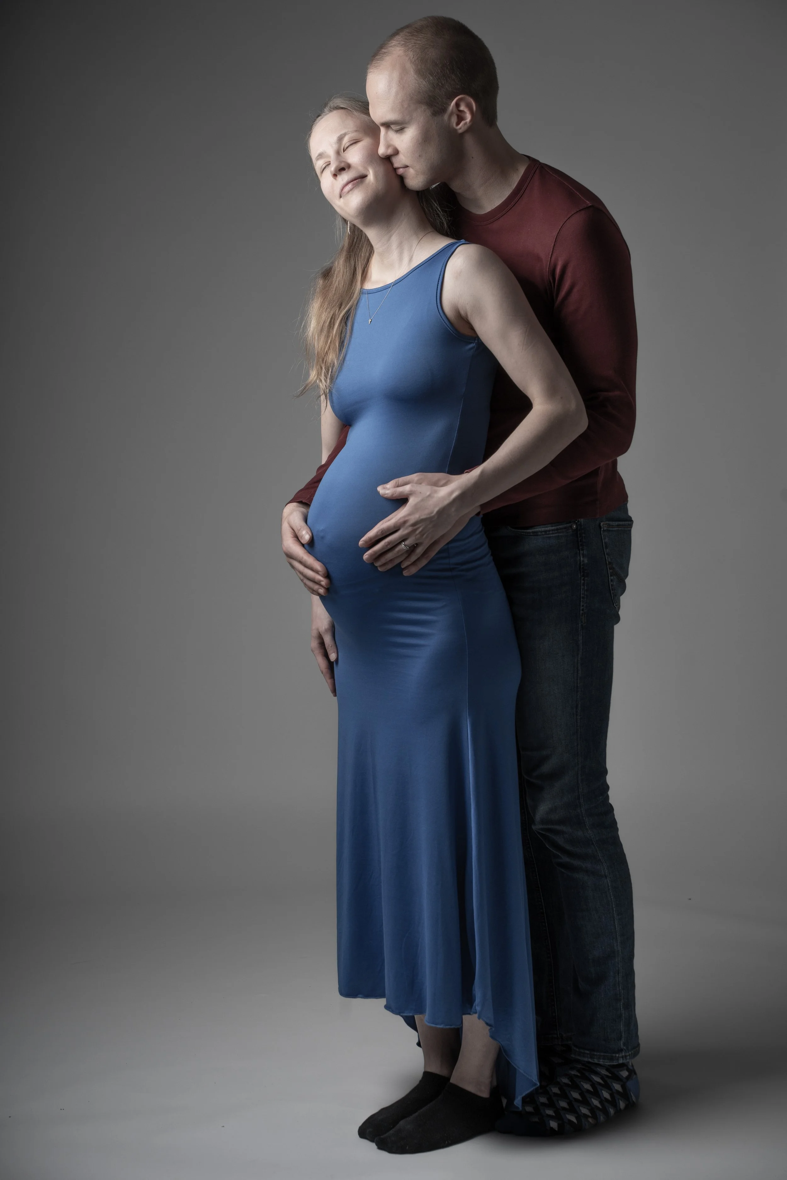 A pregnant woman in a blue dress with a man standing behind her, both touching her pregnant belly, in a studio with a gray background.