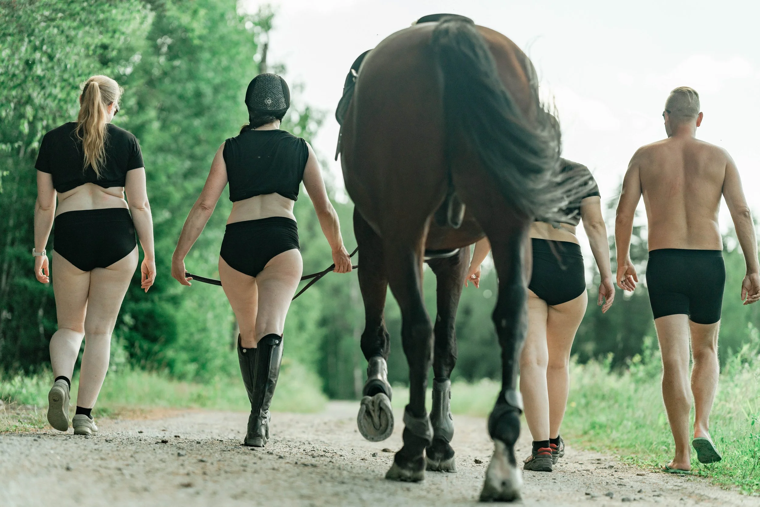 Four people and a horse walking on a dirt trail in a wooded area, wearing minimal clothing.