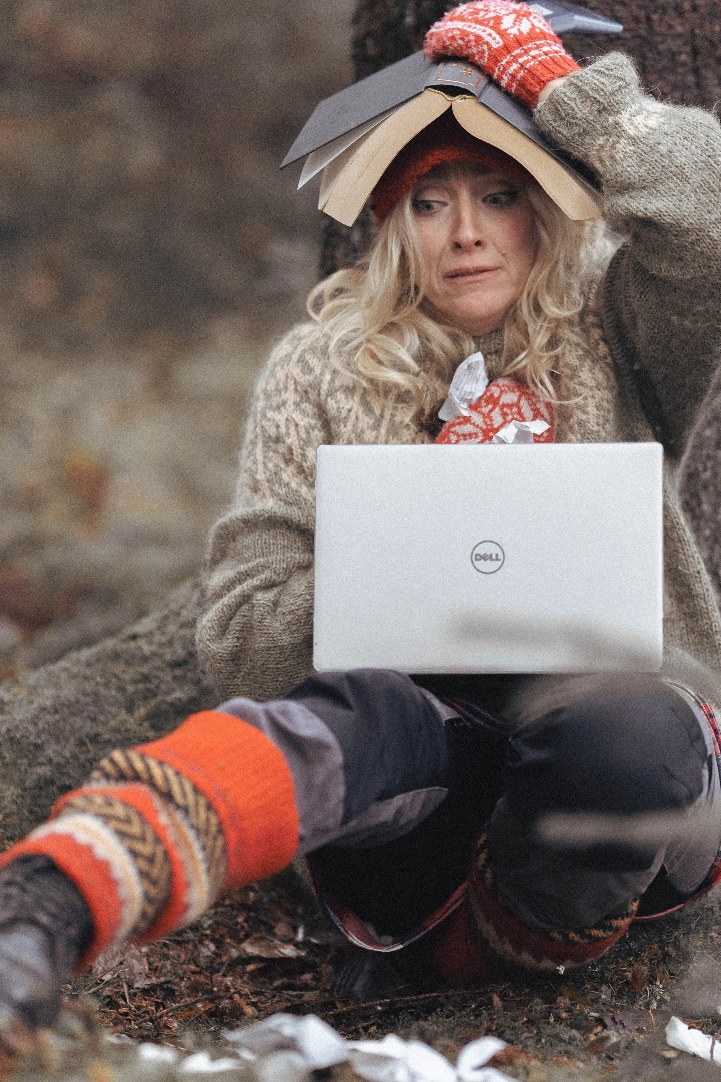 A woman sitting outdoors on the ground, holding a laptop on her lap, with a stack of open books on her head, appearing confused or overwhelmed.