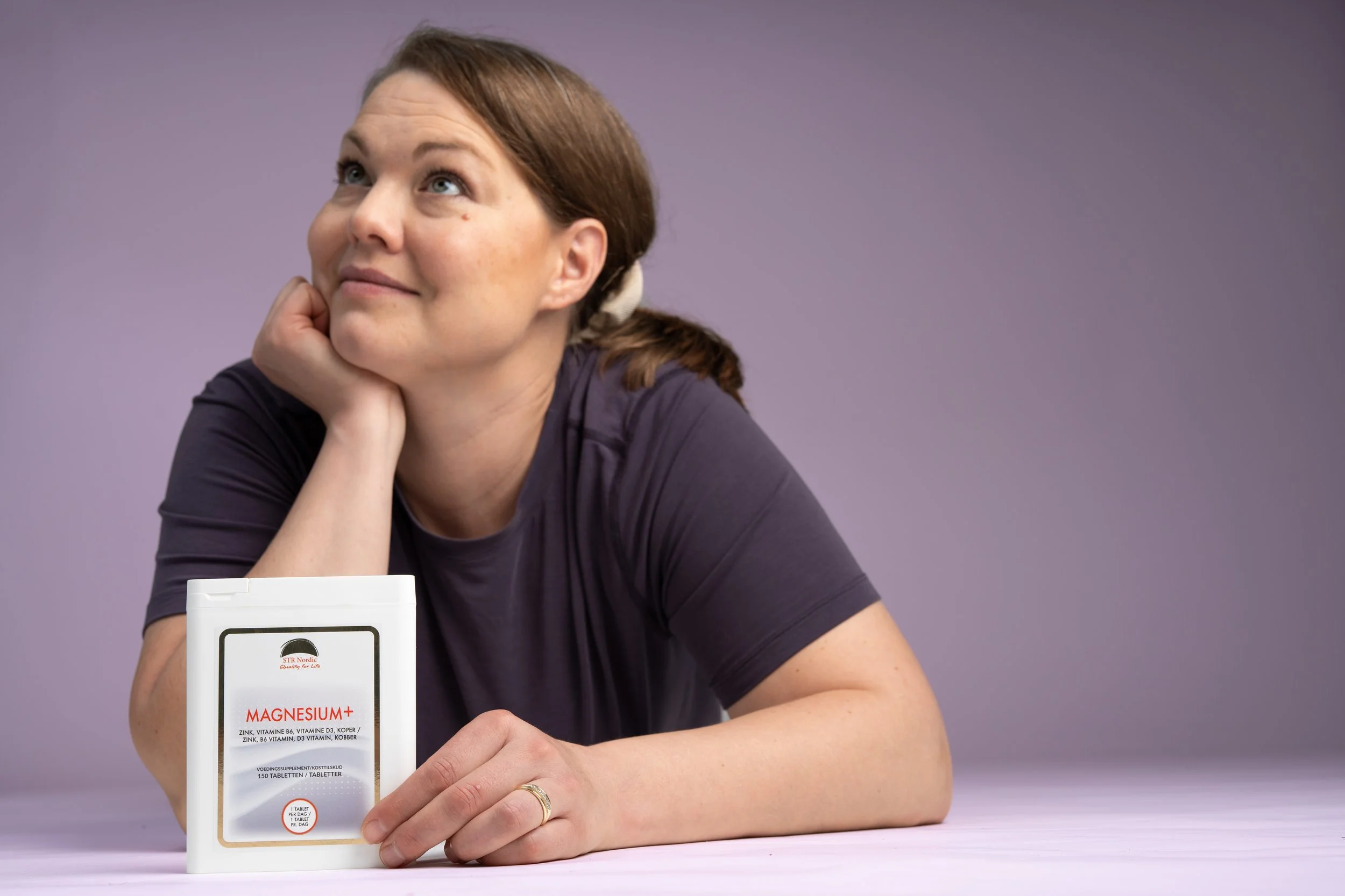 A woman with brown hair tied back, wearing a dark shirt, resting her chin on her hand, and holding a bottle of magnesium supplements on a white table against a purple background.