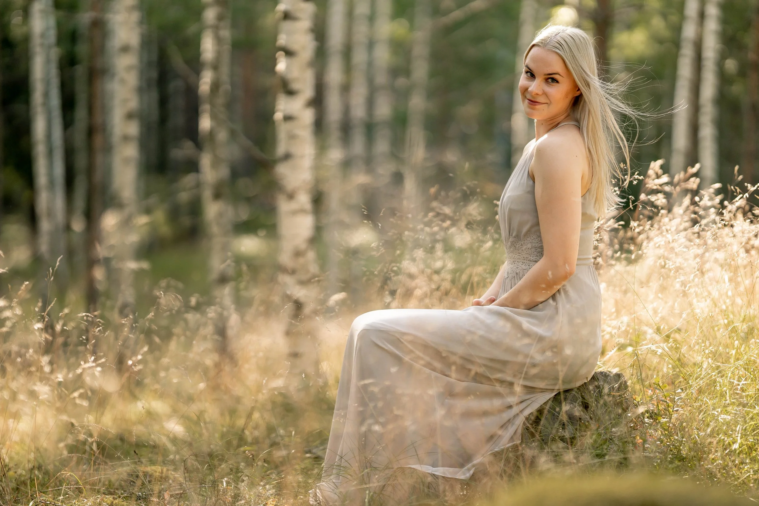 A young woman with blonde hair wearing a beige dress sitting on a log in a forest with tall trees and sunlight filtering through the leaves.
