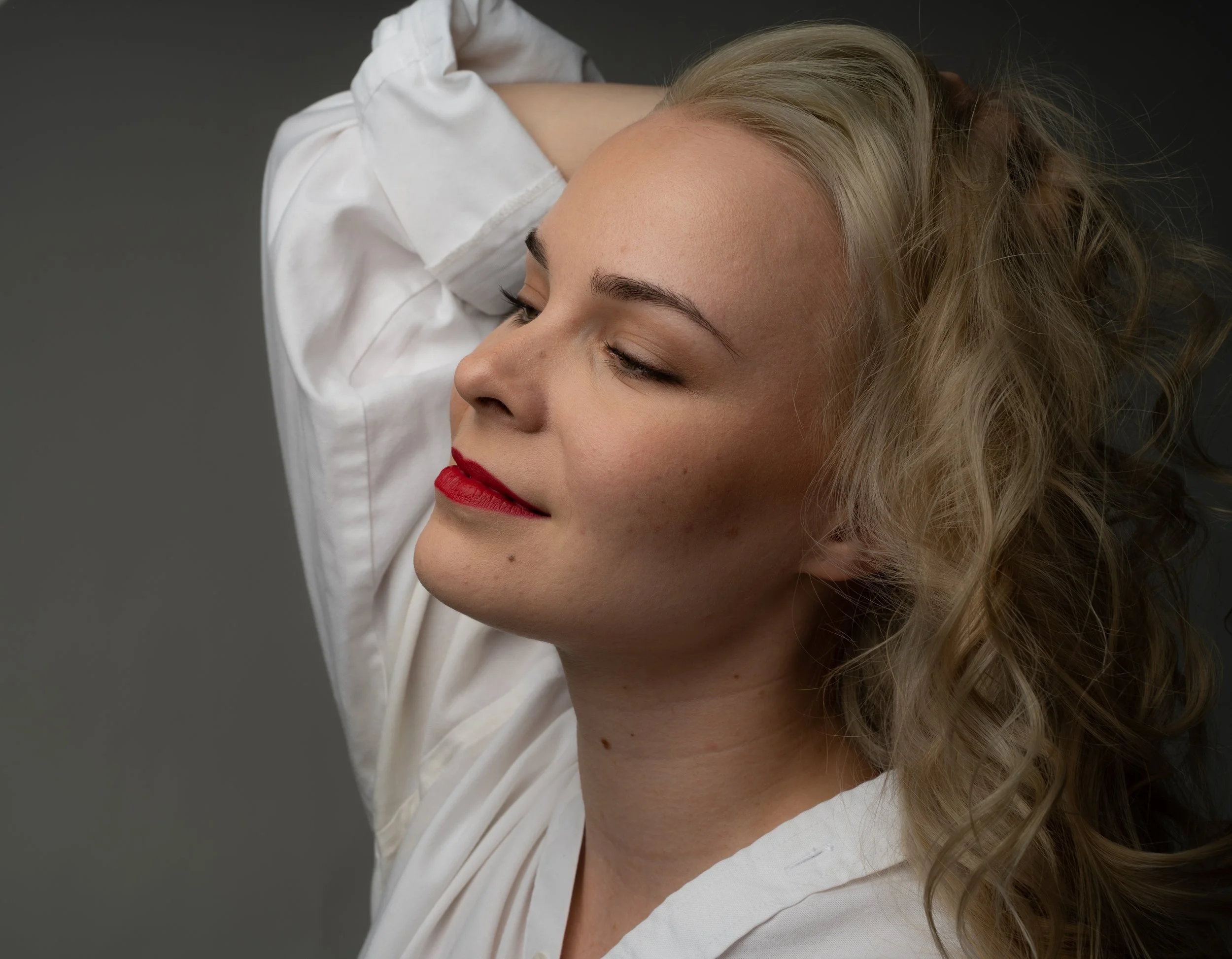 A woman with blonde, curly hair, closed eyes, and red lipstick, resting with one arm behind her head, wearing a white shirt against a neutral background.