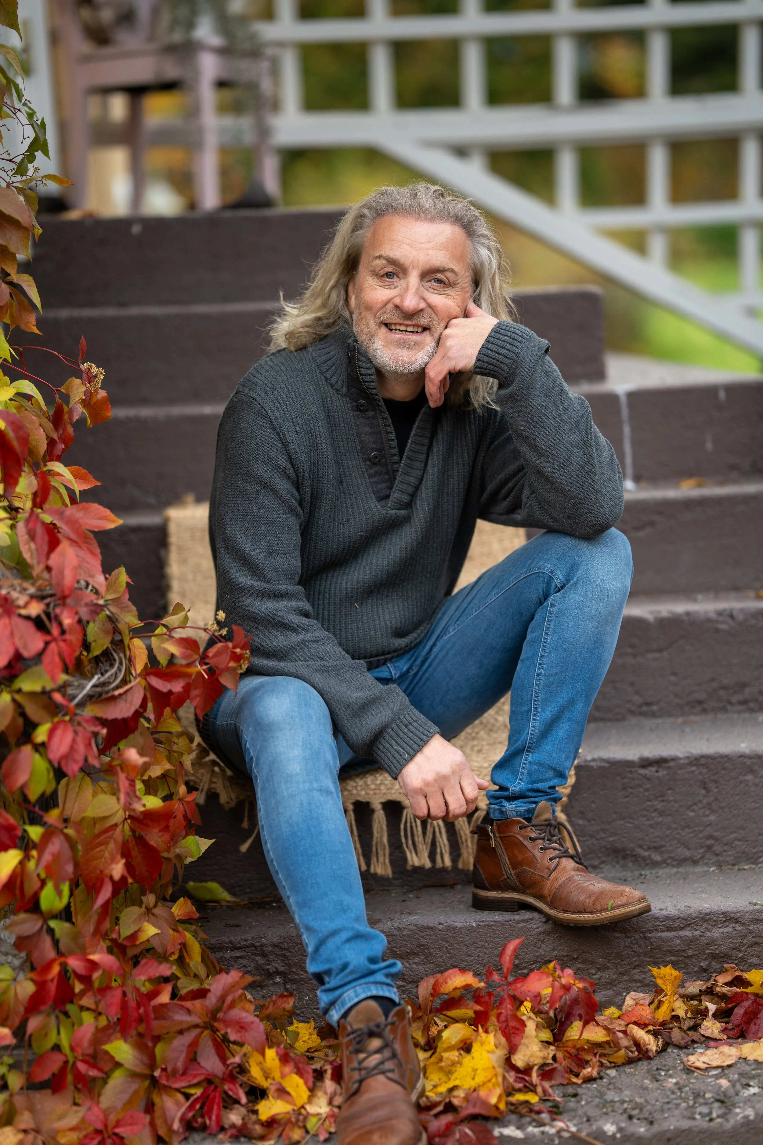 A man with long gray hair and a beard sitting on outdoor stairs surrounded by fall leaves, smiling and resting his head on his hand.