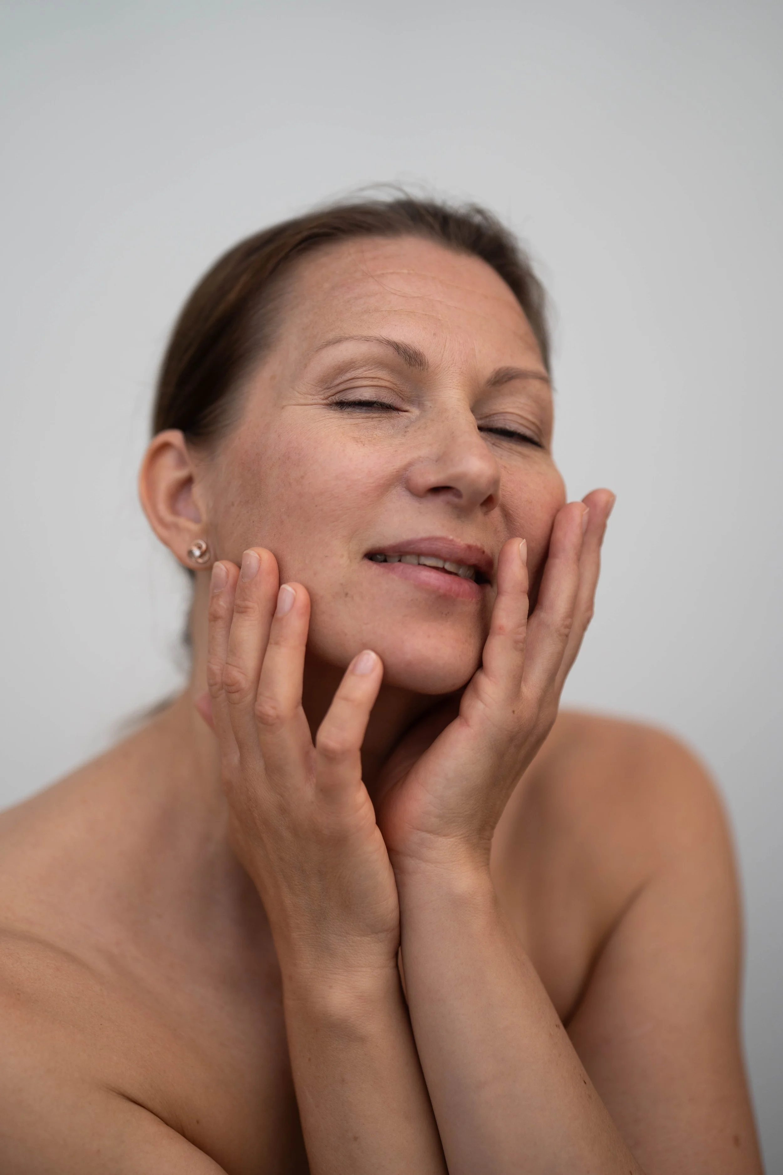 A woman with short brown hair and earrings gently touching her face with her eyes closed and a relaxed expression.