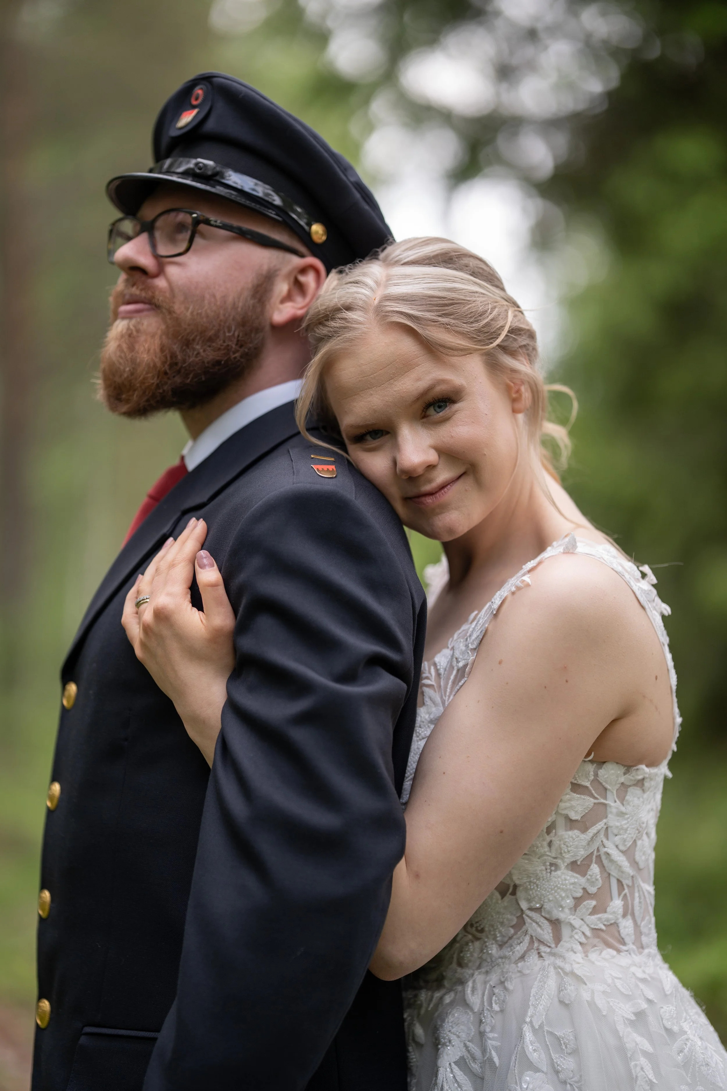 A couple, a man in a formal military uniform and a woman in a white lace dress, standing close together outdoors with greenery in the background. The woman is resting her head on the man's shoulder and smiling at the camera.