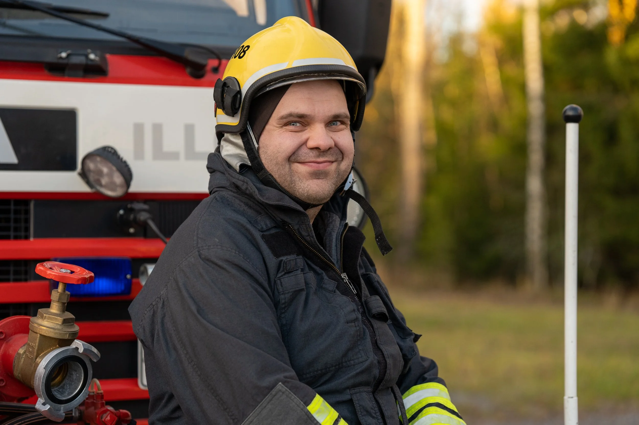 Male firefighter wearing a yellow helmet and black protective gear smiling outdoors near a fire truck.