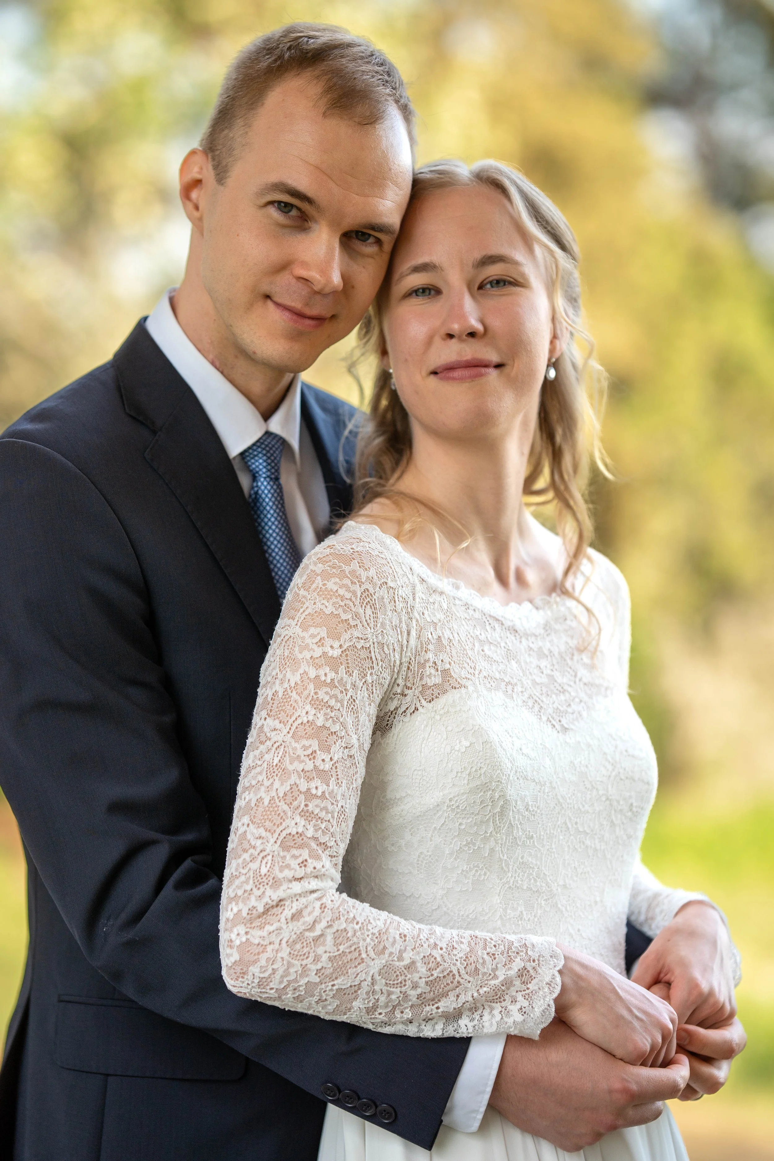 A couple in wedding attire outdoors during autumn, with the man in a dark suit and the woman in a lace wedding dress, standing close together.