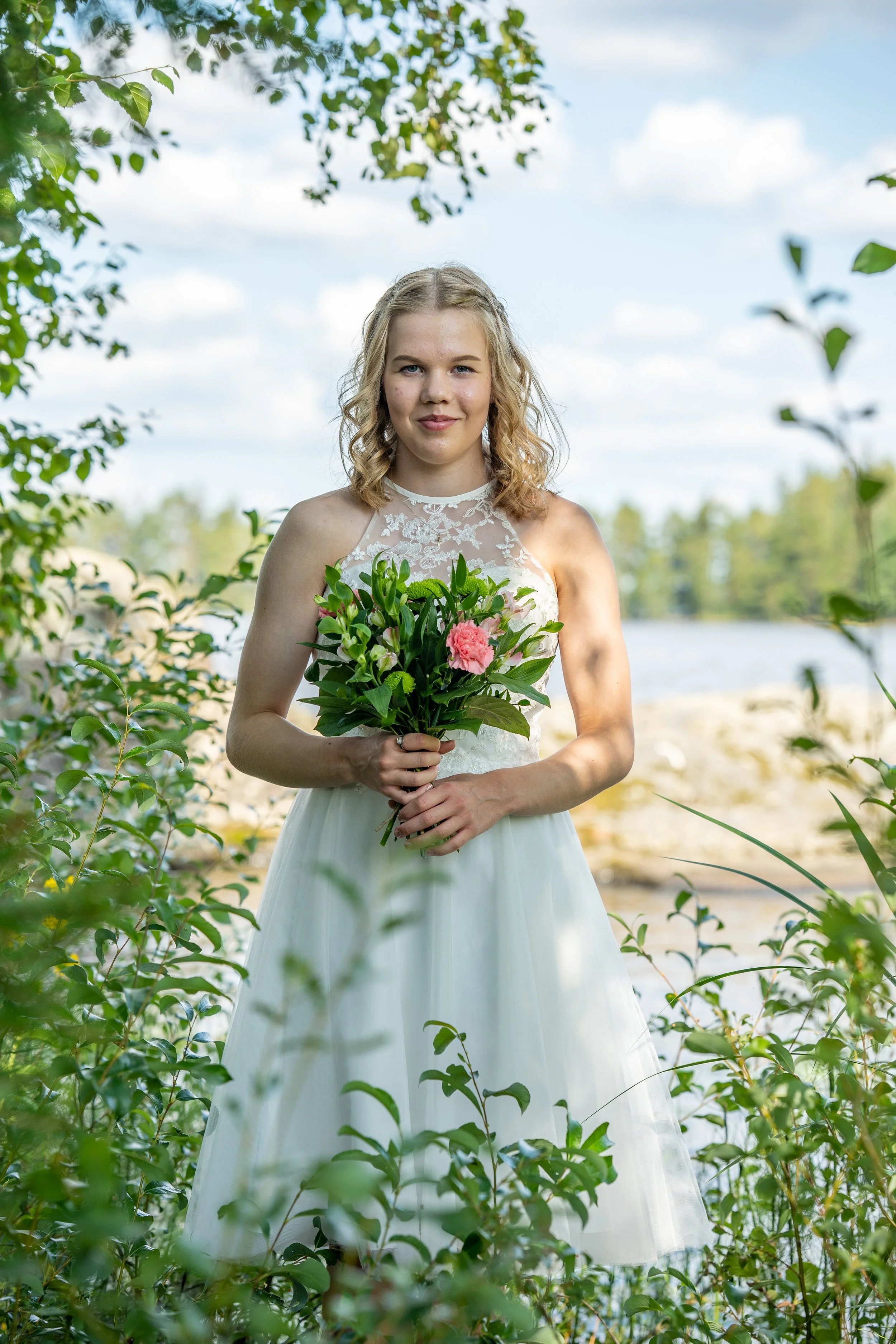 A young woman in a white dress holding a bouquet of pink and white flowers outdoors near water and green foliage.