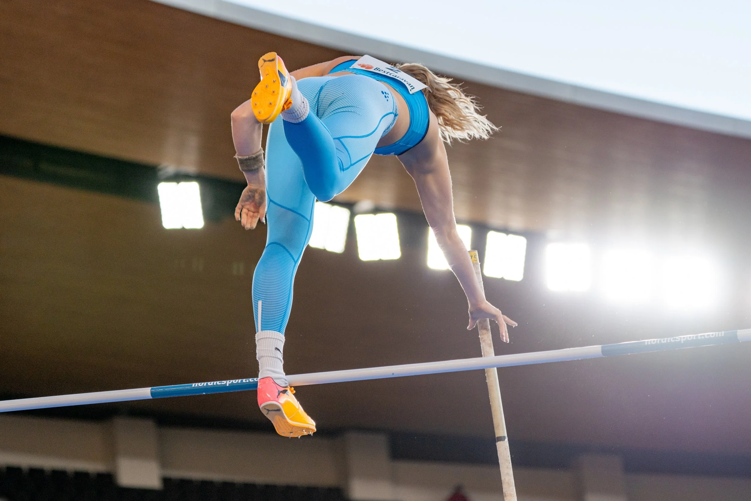 A female pole vaulter in blue athletic gear clearing a high bar during a competition inside an indoor stadium.