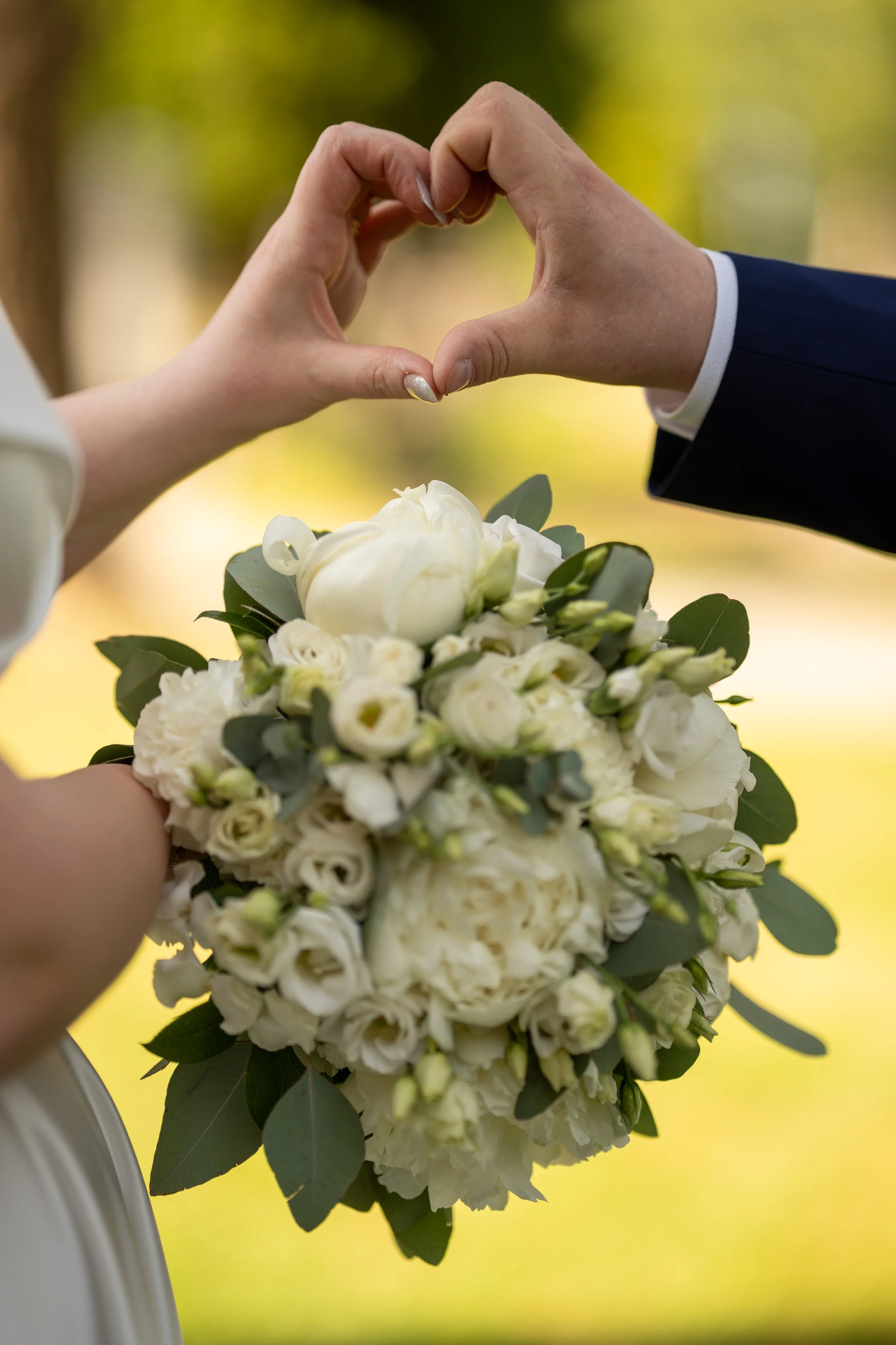 Two people forming a heart shape with their hands over a white flower bouquet during a wedding.