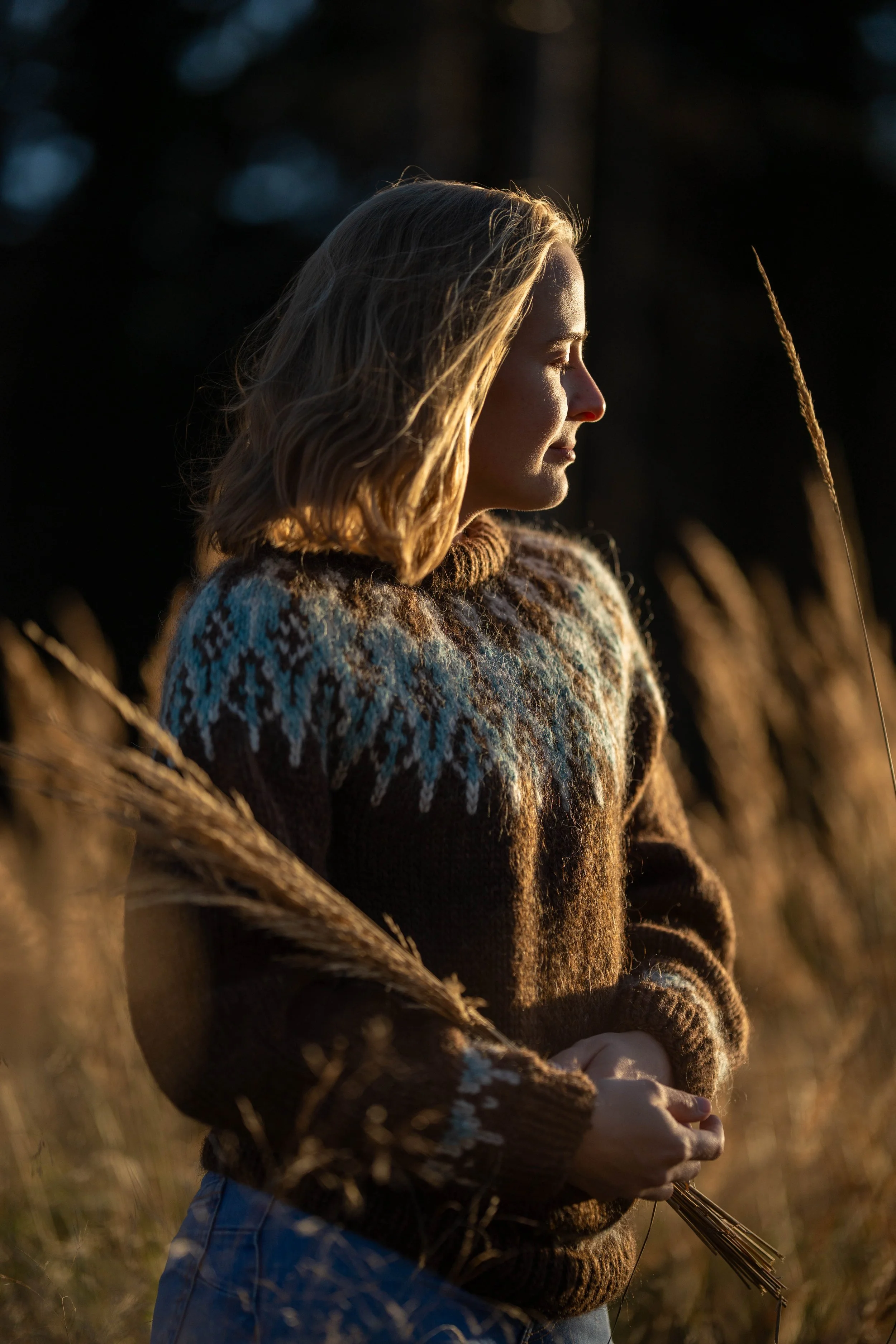 A woman with shoulder-length hair stands in a field of tall grass during sunset, facing sideways with her eyes closed.