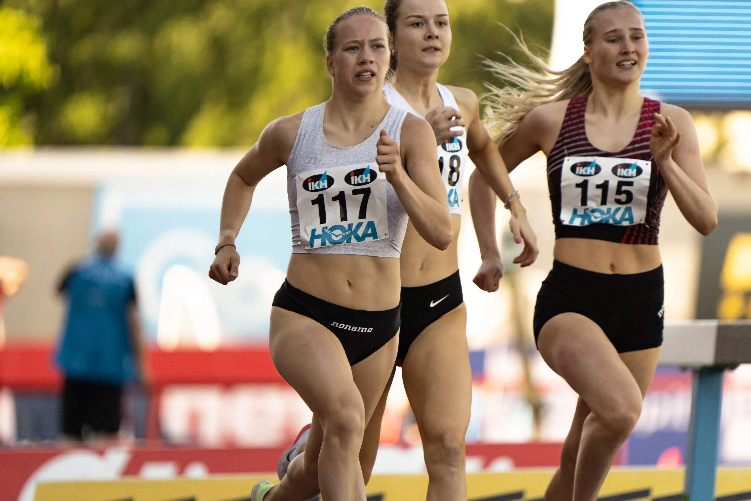 Three women running in a race, wearing athletic outfits and race bibs with numbers 117, 118, and 115.