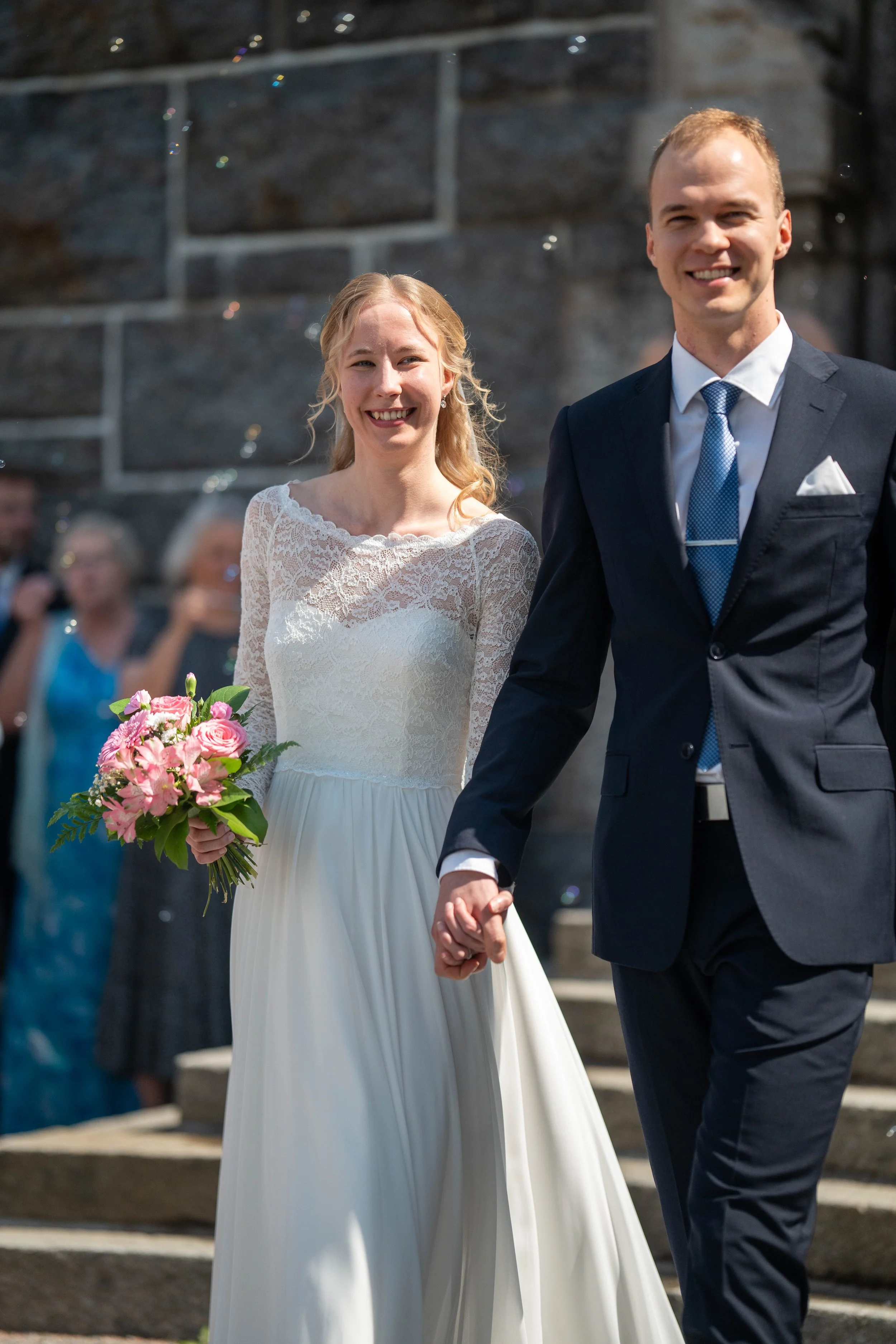 A bride and groom holding hands, walking outdoors on their wedding day, with wedding guests in the background.