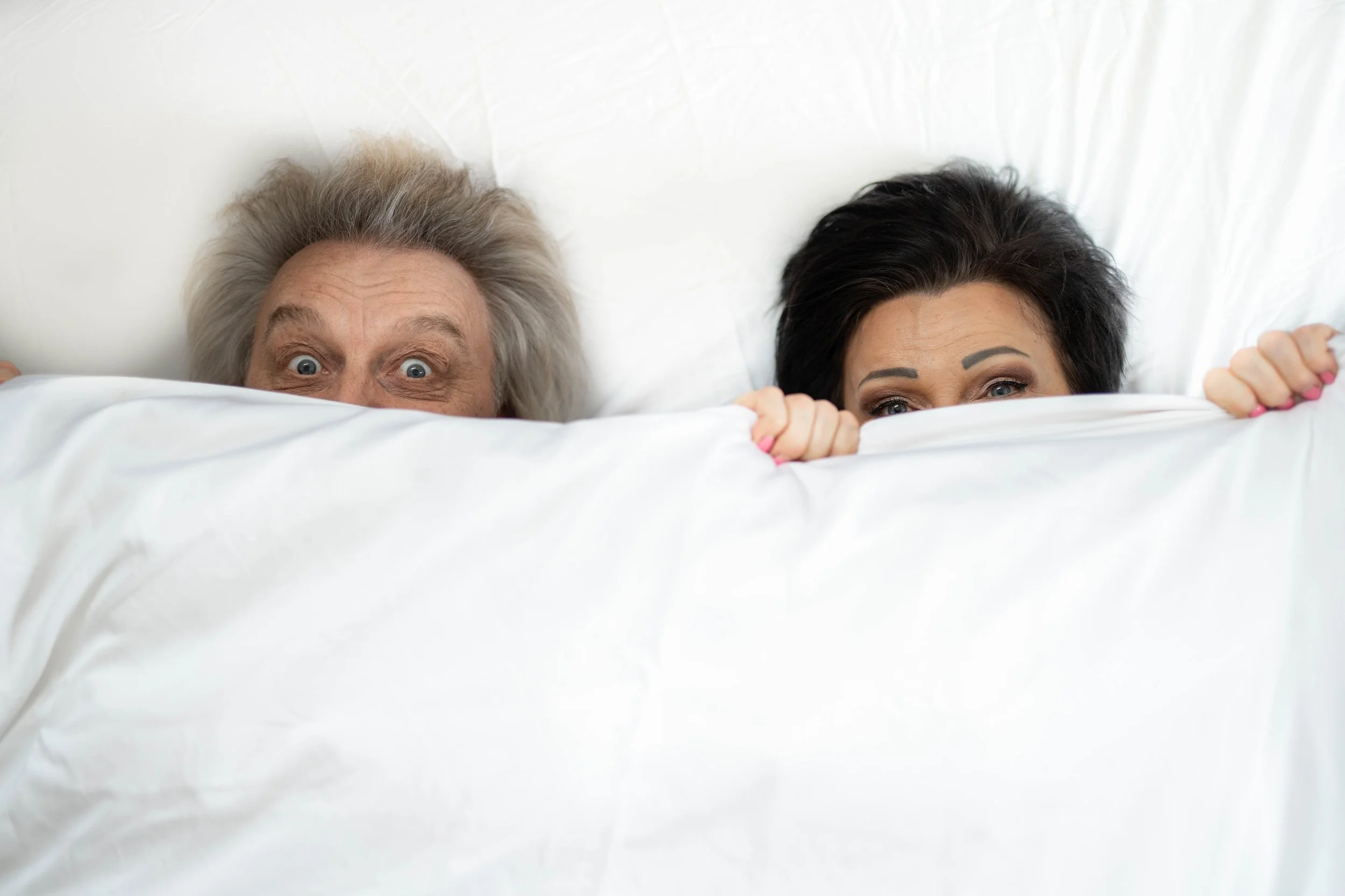 Two people hiding under a white blanket in bed, with only their eyes and part of their faces visible, one with gray hair and one with dark hair, looking surprised or playful.